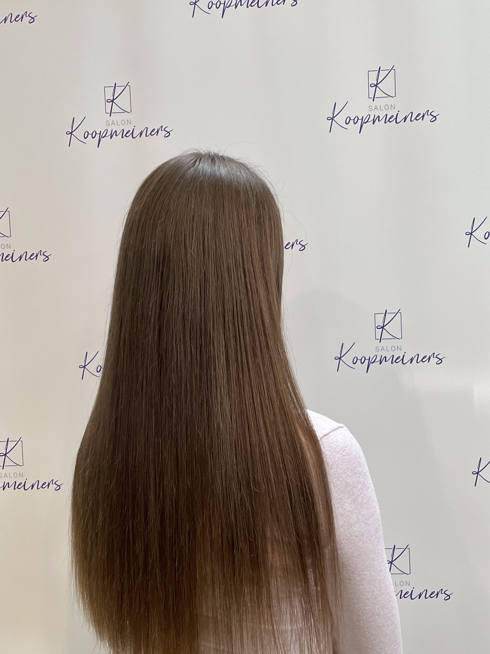 Frau mit langem, braunem, gewelltem Haar, mit dem Gesicht abgewandt, in einem Studio mit einer Wand mit Logomuster.