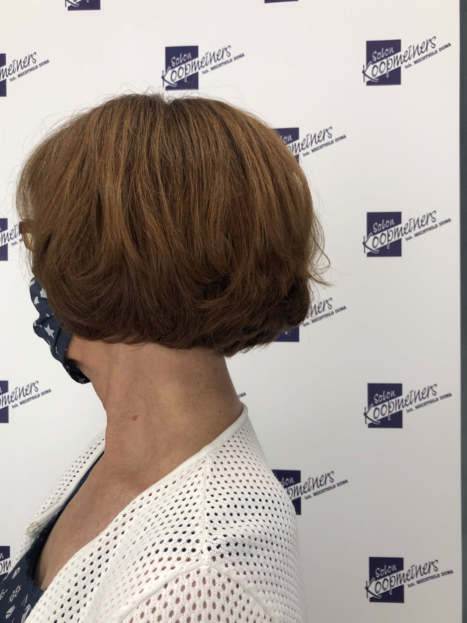 Frau mit kurzen, braunen, lockigen Haaren, trägt eine Gesichtsmaske und einen weißen Pullover, Profilansicht.