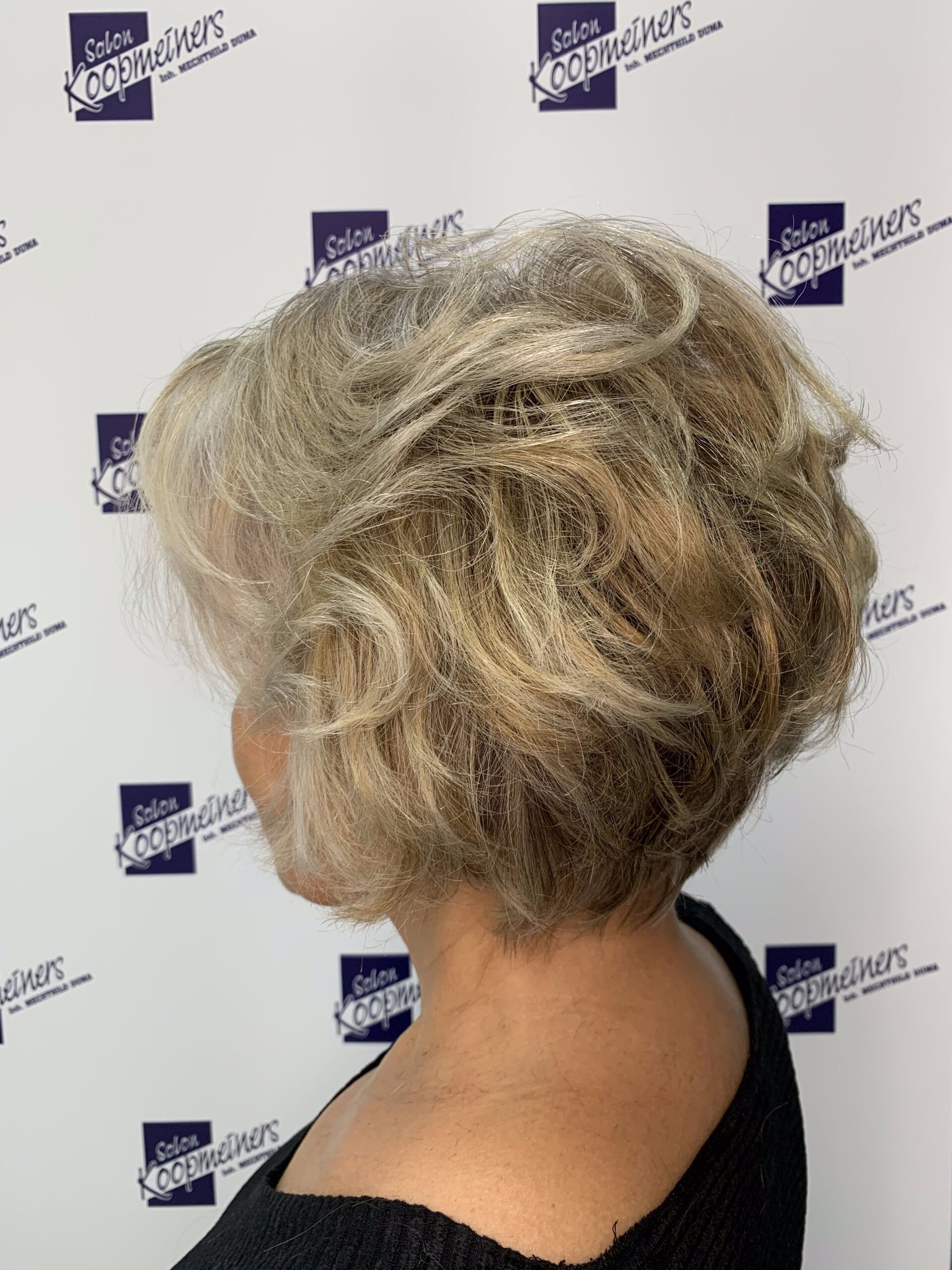 Frau mit kurzem, welligem, grauem und blondem Haar, nach links blickend, trägt ein schwarzes Oberteil.