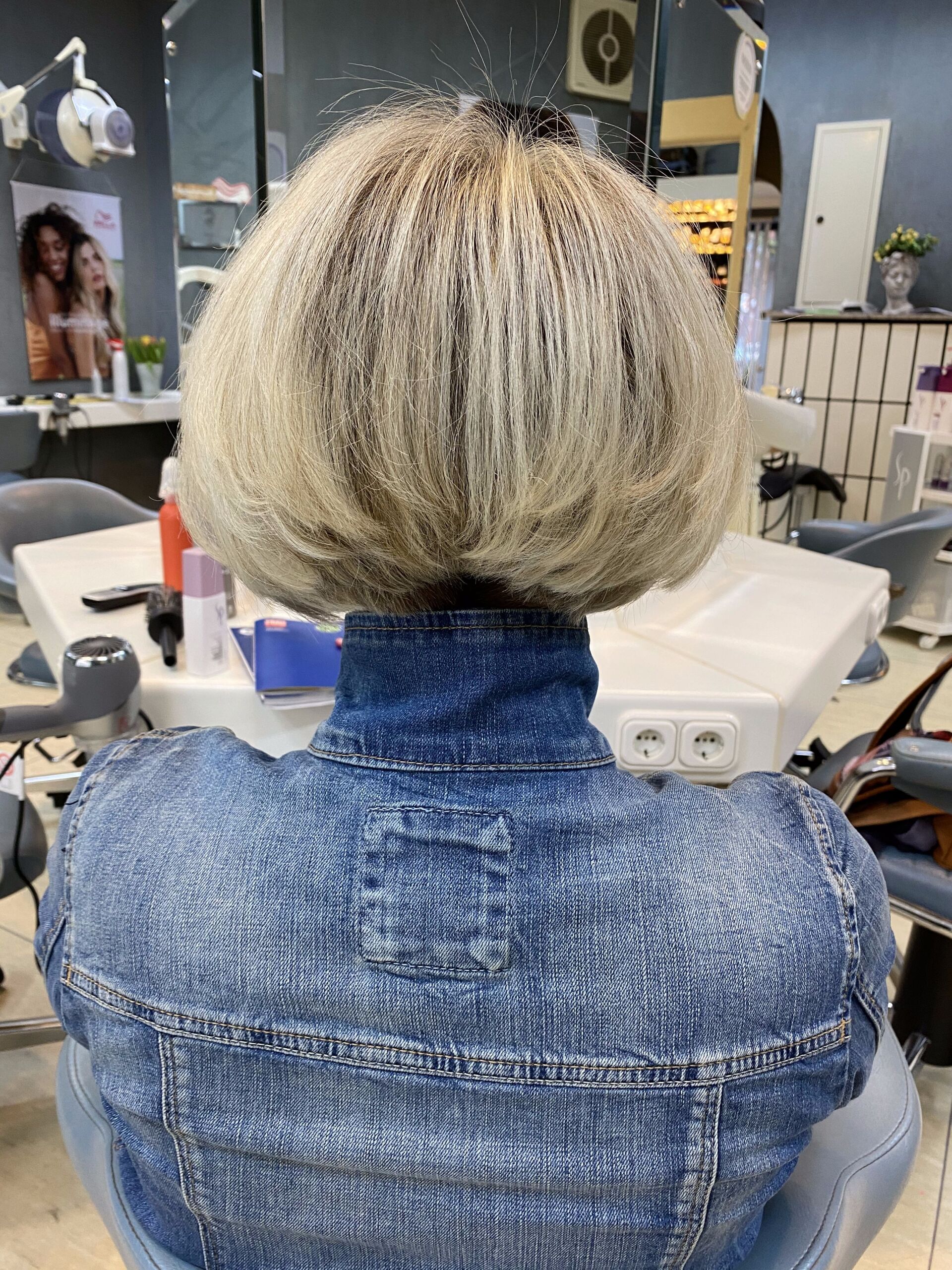 Frau mit blonder Bobfrisur, trägt eine blaue Jeansjacke, in einem Salon.