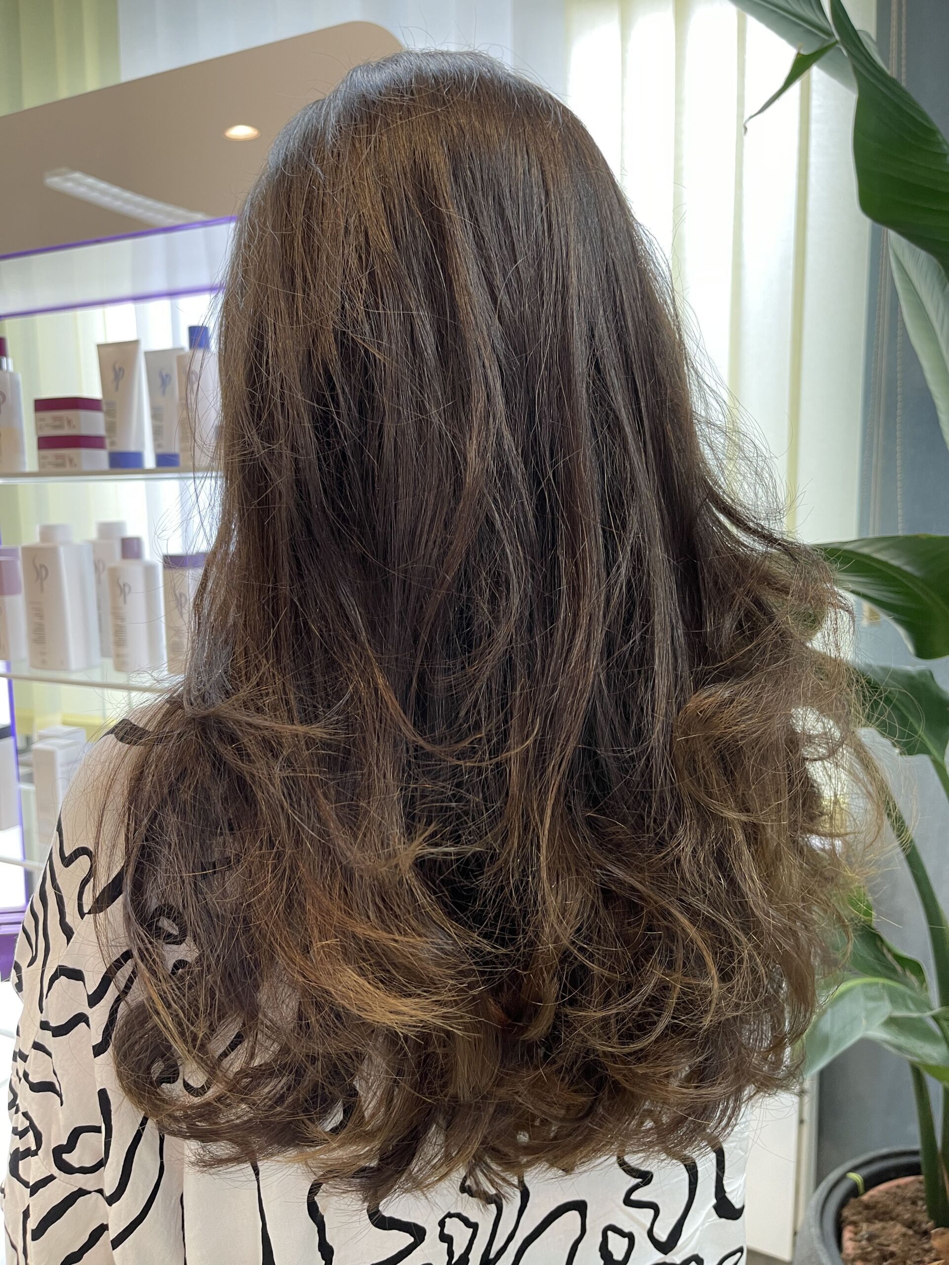 Frau mit dunklem, welligem Haar in einem Salon, von hinten gesehen.