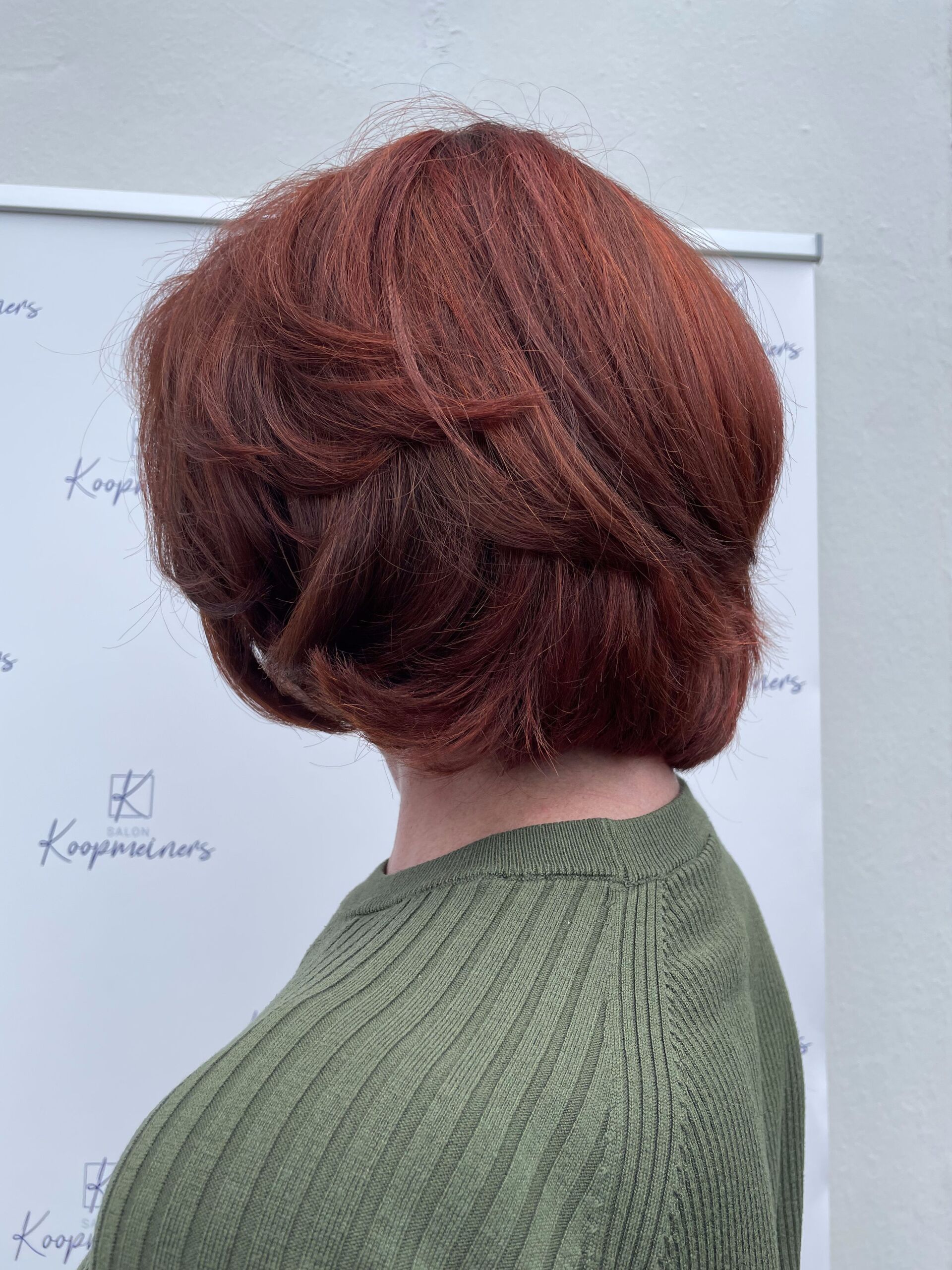 Frau mit gestuftem rotem Haar und grünem Pullover, zur Seite gedreht.