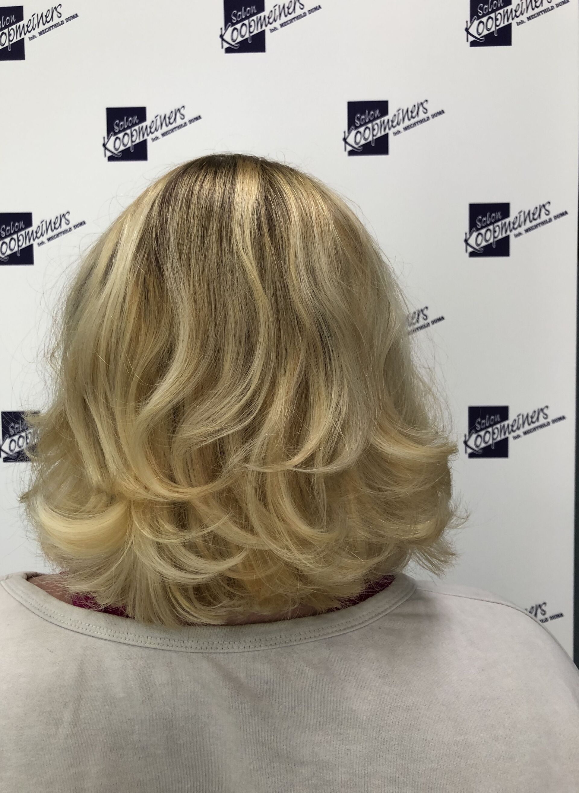 Blonder, gestufter Bob-Haarschnitt. Dunkle Haarwurzeln, Rückansicht. Heller Pullover. Weißer Hintergrund mit Logos.