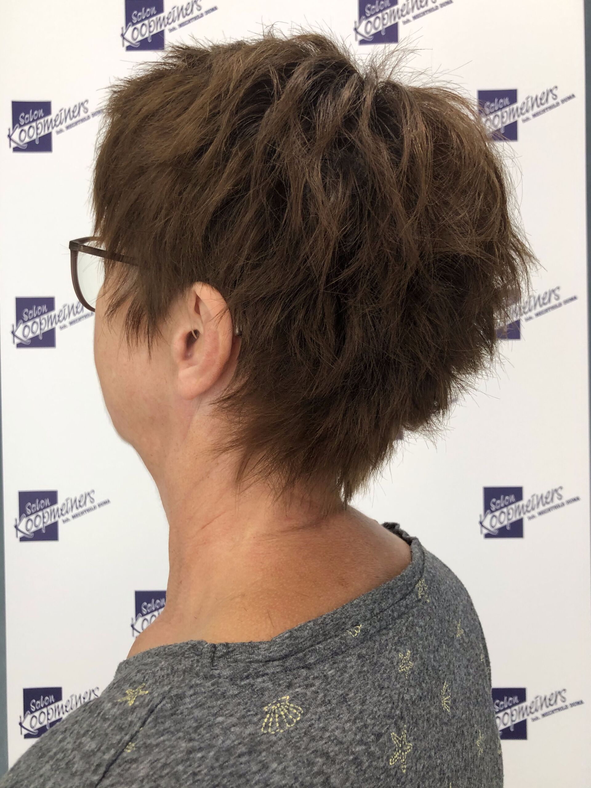 Frau mit kurzem, strukturiertem braunem Haar, Brille und grauem Pullover.