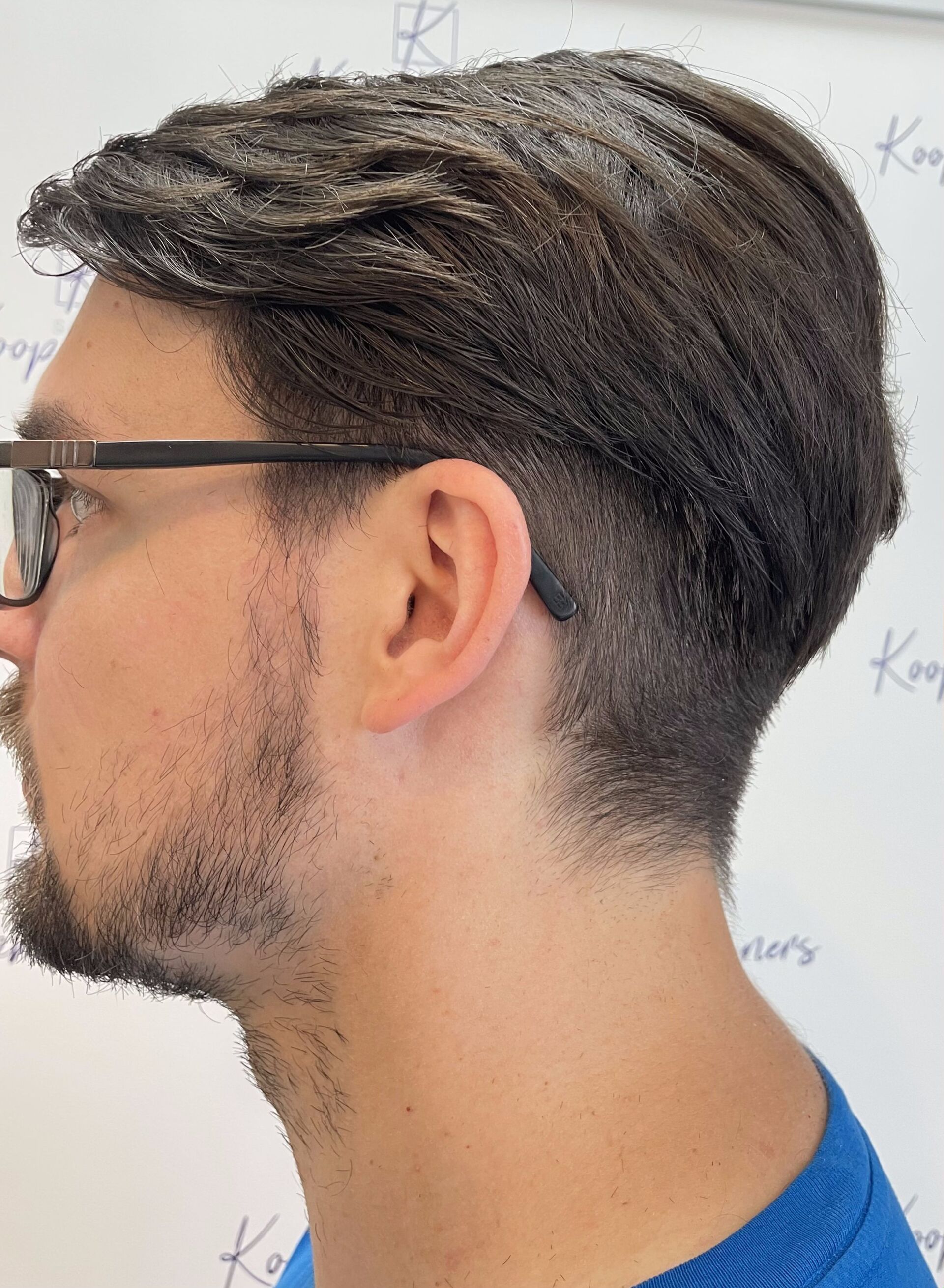 Mann mit dunklem Haar und Undercut-Frisur, Brille und blauem Hemd, Blick nach links.