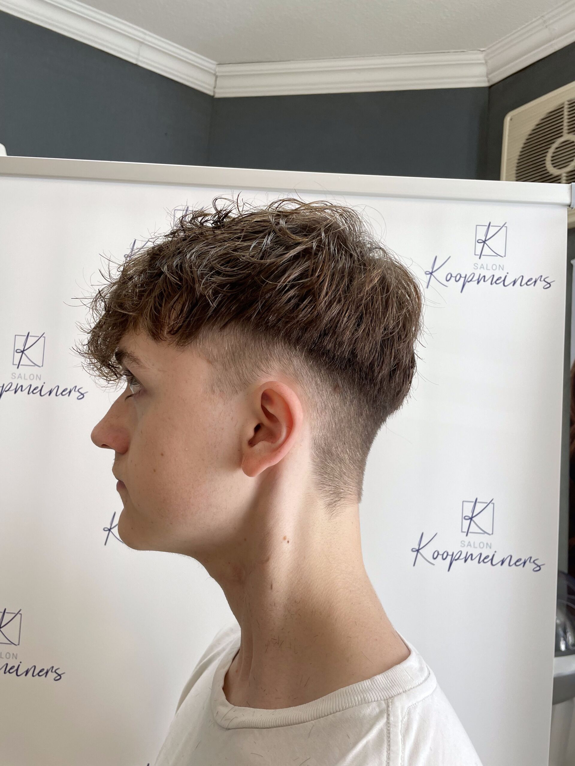 Ein junger Mann mit verblasstem Haarschnitt. Lockiges braunes Haar oben. Weißer Hintergrund mit Logos.