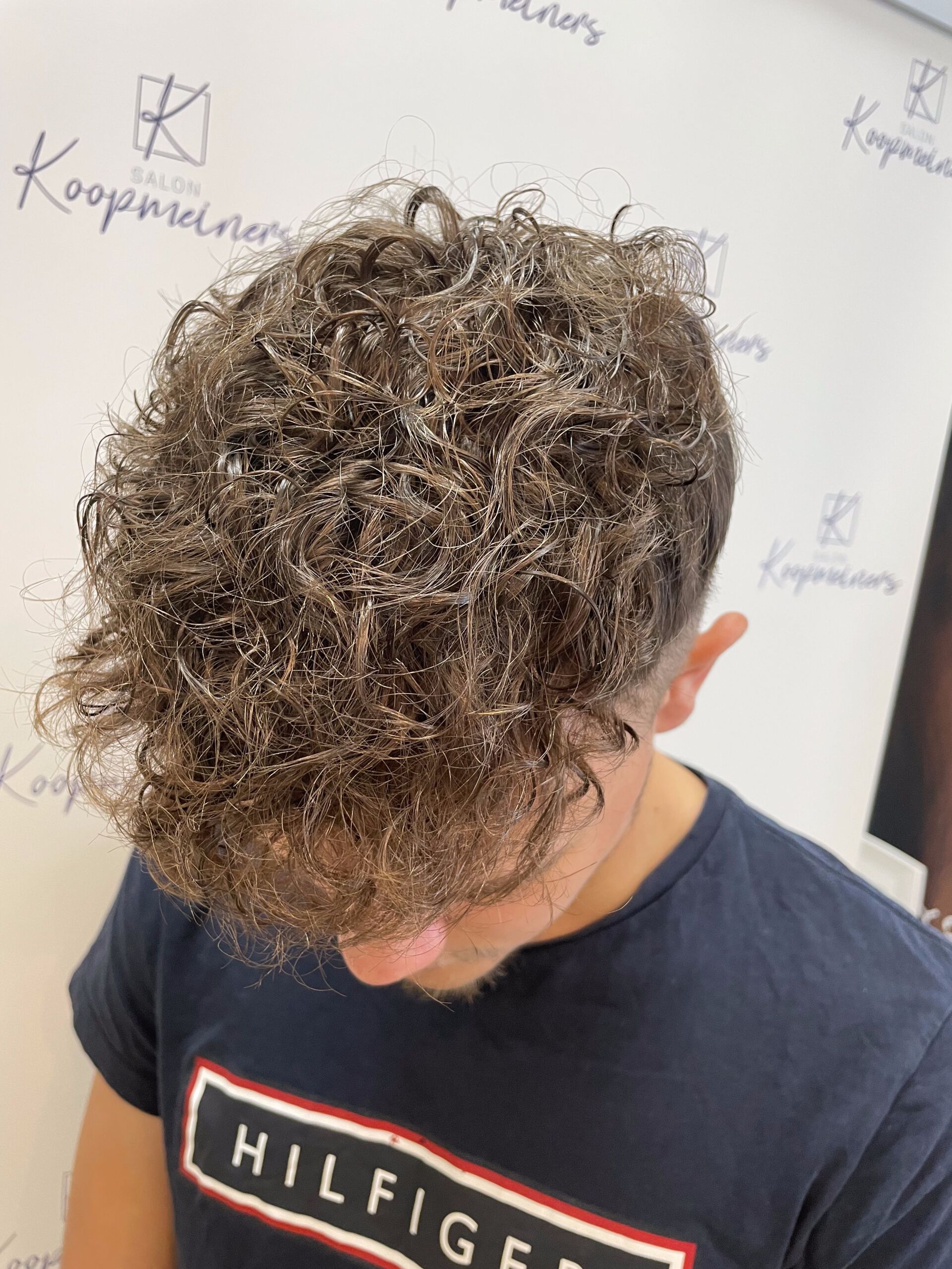 Mann mit lockigem braunem Haar, trägt ein marineblaues Hemd und blickt nach unten. Weiße Wand mit Logo im Hintergrund.