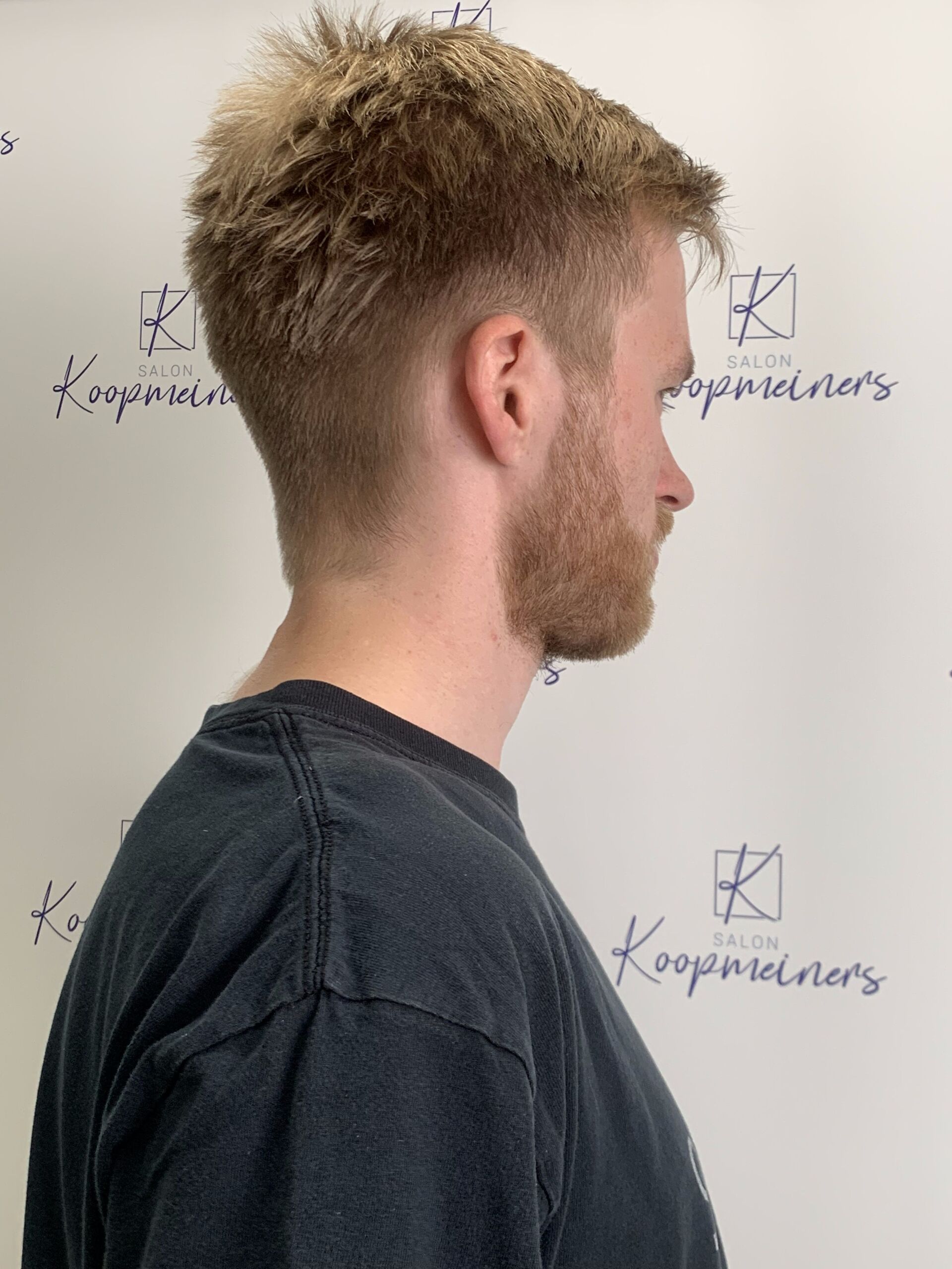 Mann mit blondem Haar und Bart, trägt ein schwarzes Hemd, Profilansicht, in einem Salon.