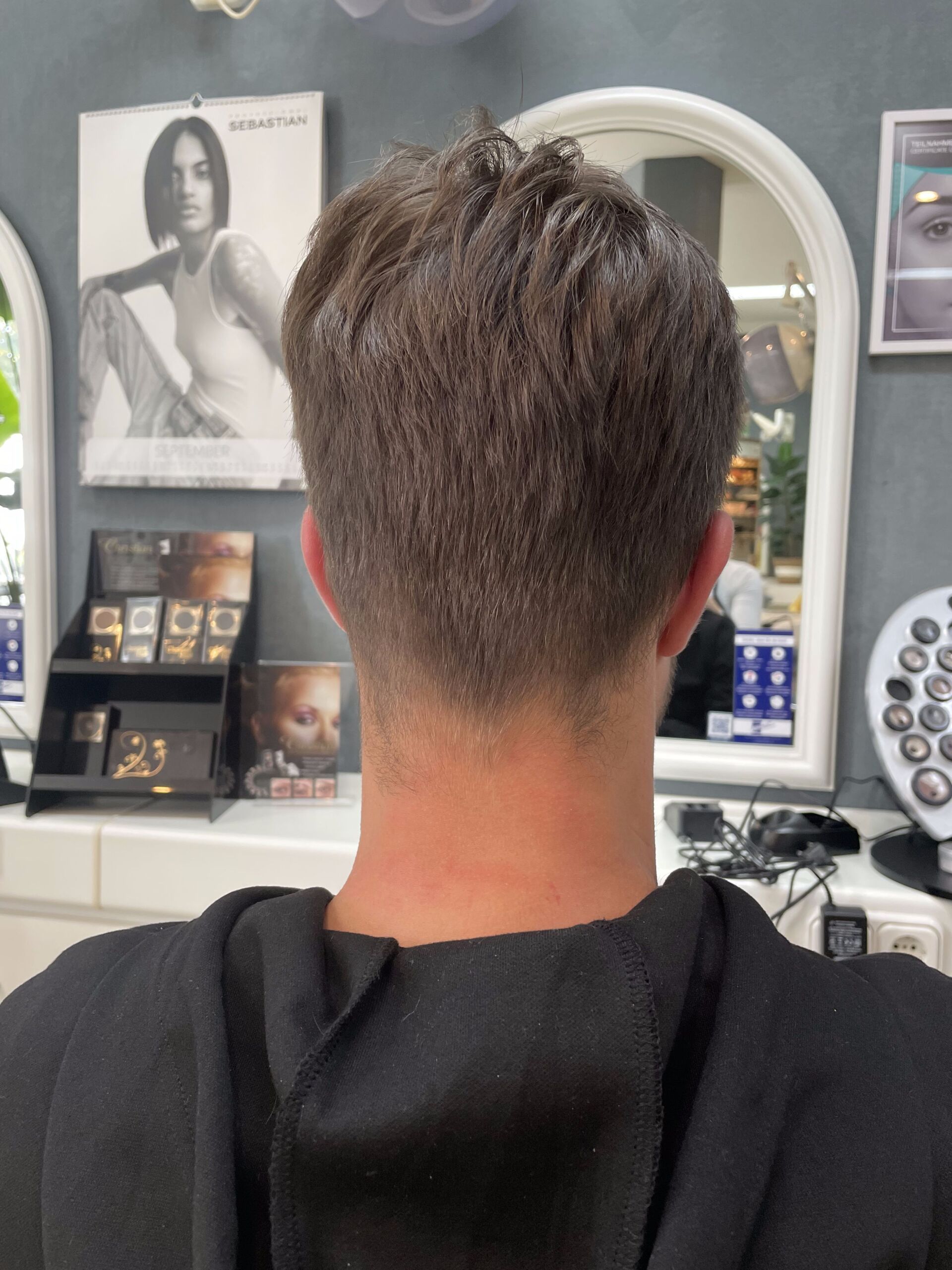 Mann mit kurzen braunen Haaren in einem Salon, von hinten gesehen. Bekleidet mit einem schwarzen Kapuzenshirt.