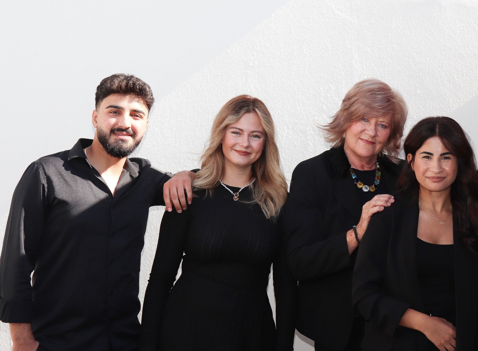 Vier Menschen unterschiedlicher Herkunft stehen vor einer weißen Wand, alle in schwarzer Kleidung und lächelnd.