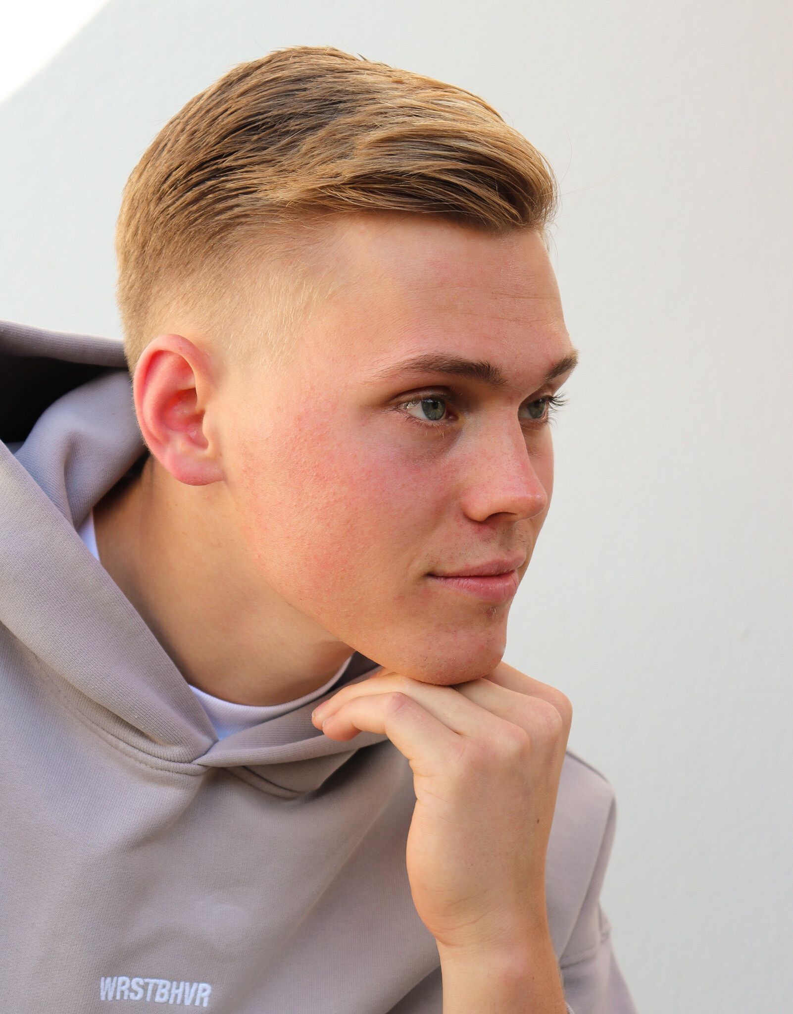 Junger Mann mit blondem Haar, das Kinn auf die Hand gestützt, trägt einen hellgrauen Kapuzenpullover.