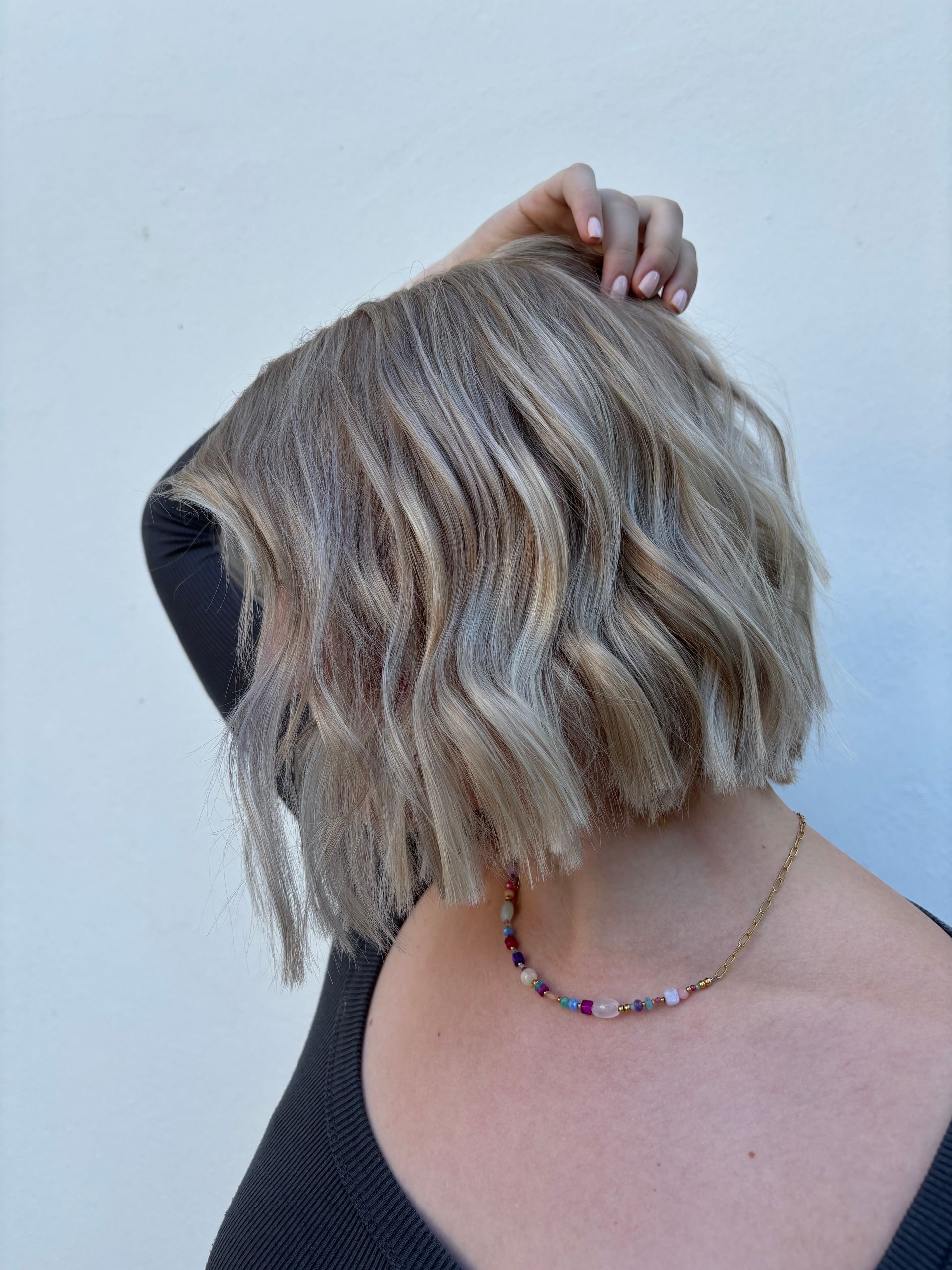 Frau mit kurzem, welligem blondem Haar berührt ihren Kopf, trägt ein schwarzes Oberteil und eine Perlenkette.