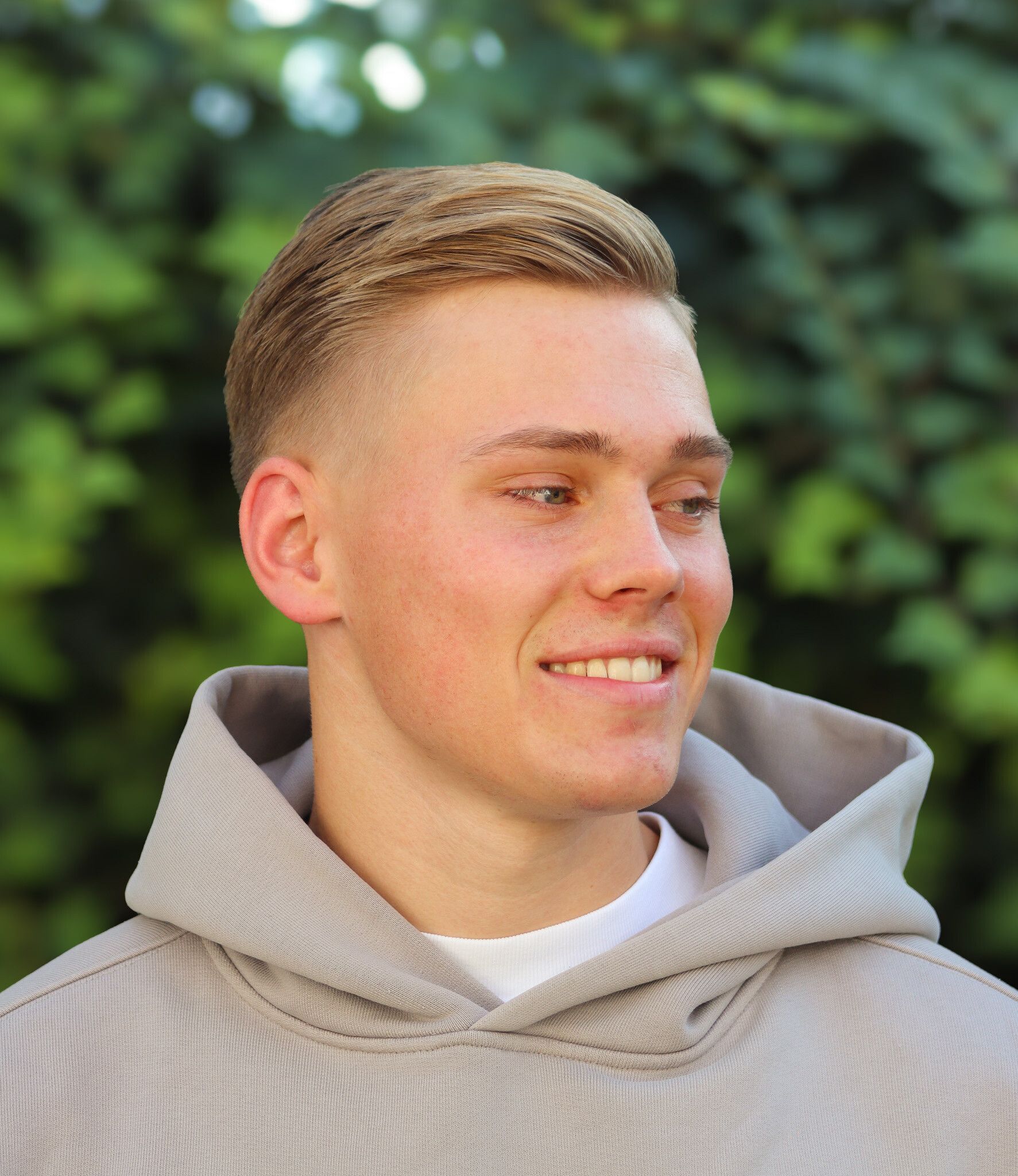 Junger Mann mit blonden Haaren lächelt und trägt im Freien einen grauen Kapuzenpullover.