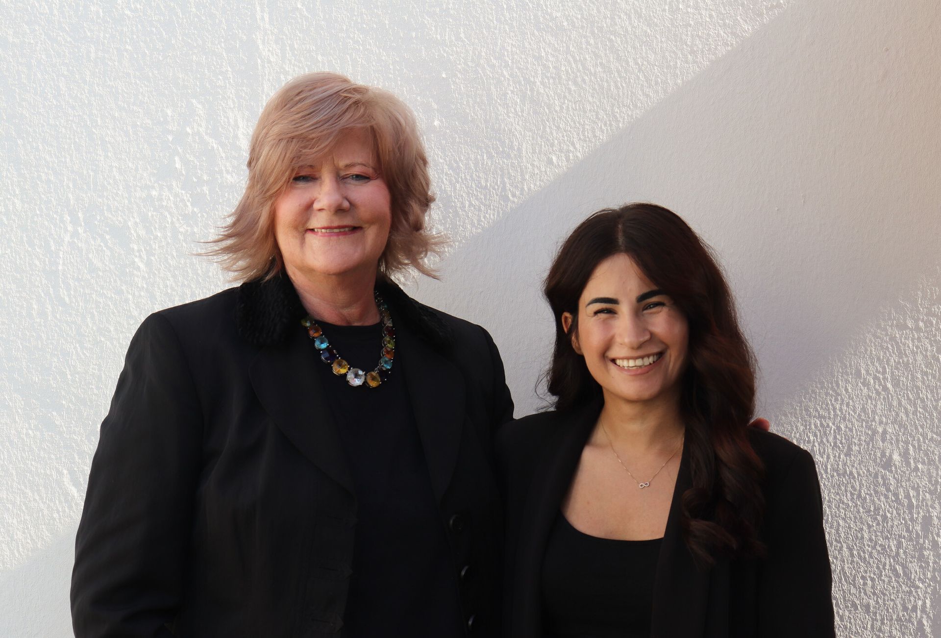 Zwei Frauen in schwarzen Blazern stehen lächelnd vor einer weißen Wand. Die eine hat blondes Haar, die andere dunkles.
