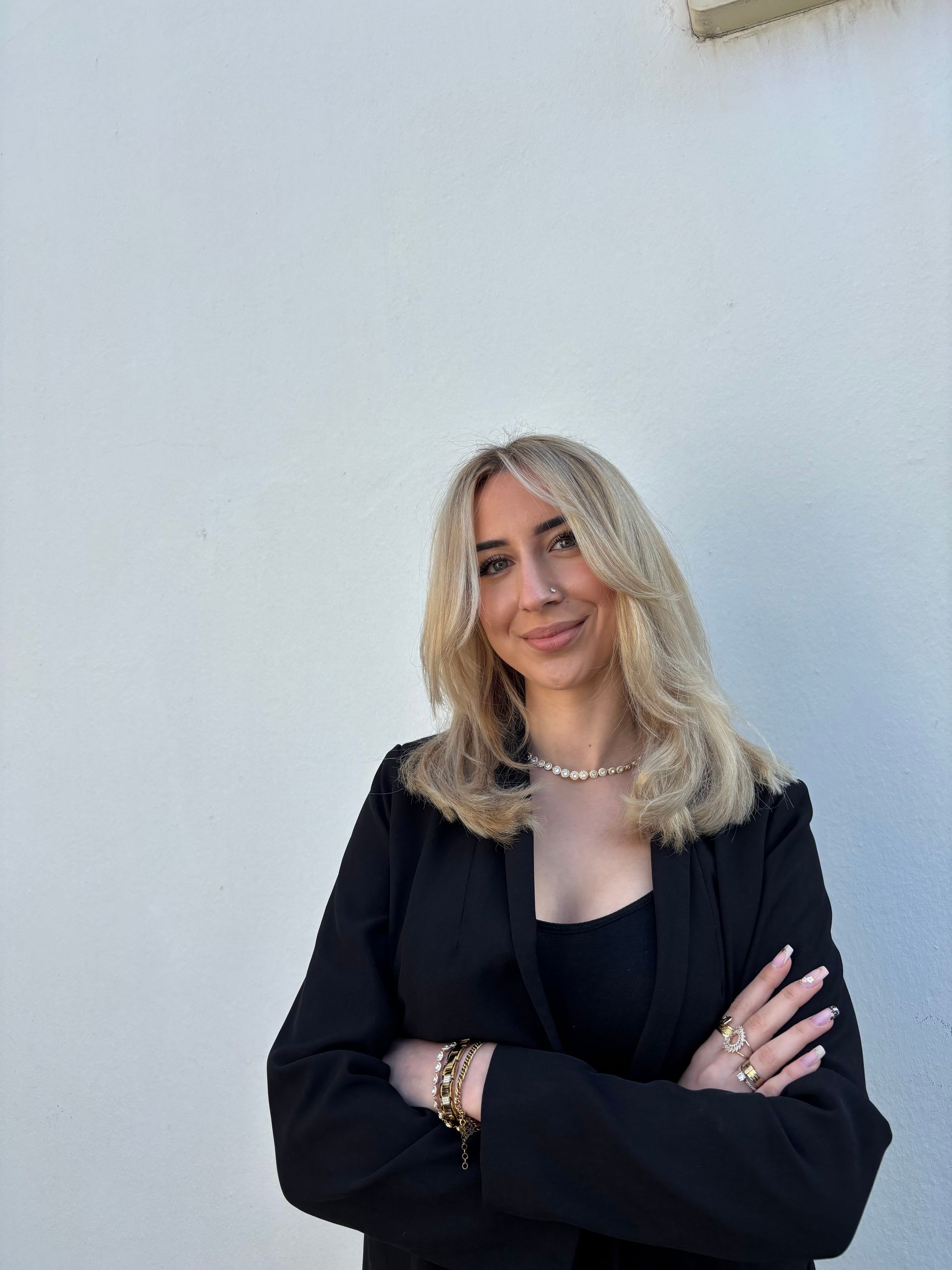 Frau mit blonden Haaren in schwarzem Blazer, Arme verschränkt, lächelt vor weißer Wand.