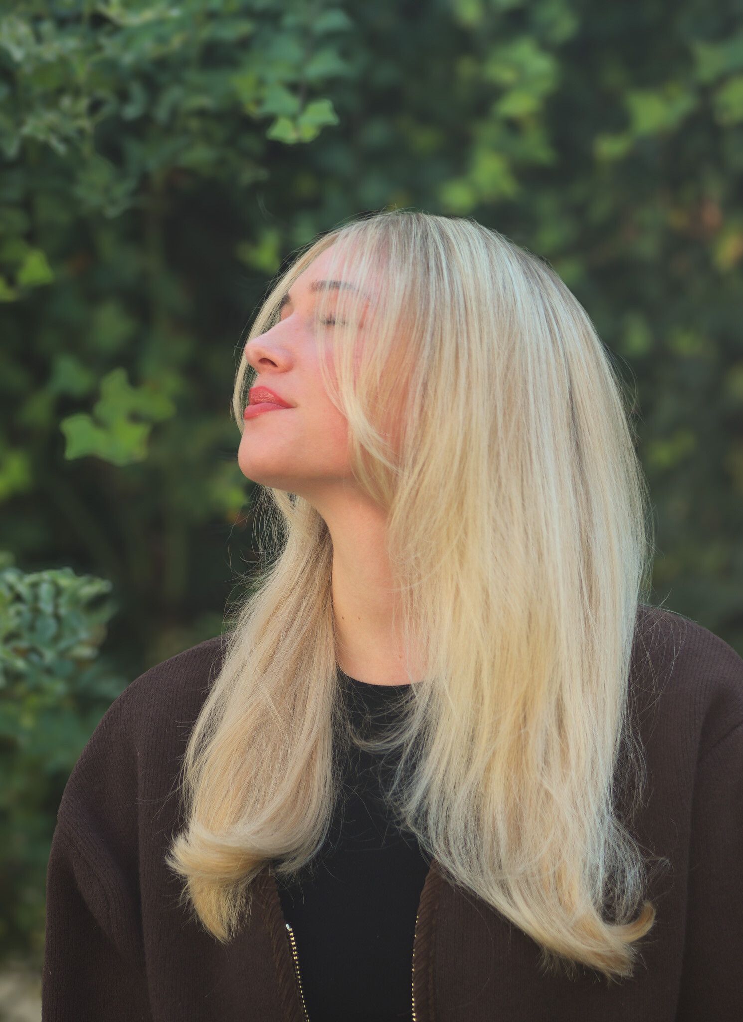 Blonde Frau mit geschlossenen Augen, geneigtem Kopf und lächelnd in der Nähe von grünem Laub