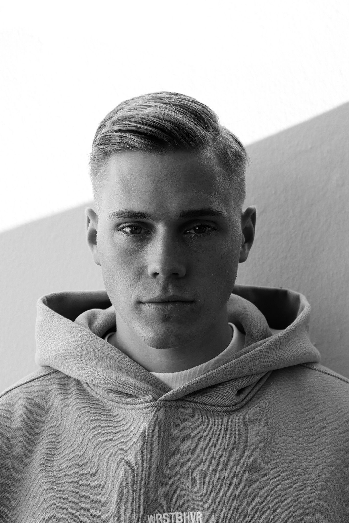 Junger Mann in einem grauen Kapuzenpullover, der vor einer hellen Wand in die Kamera blickt.