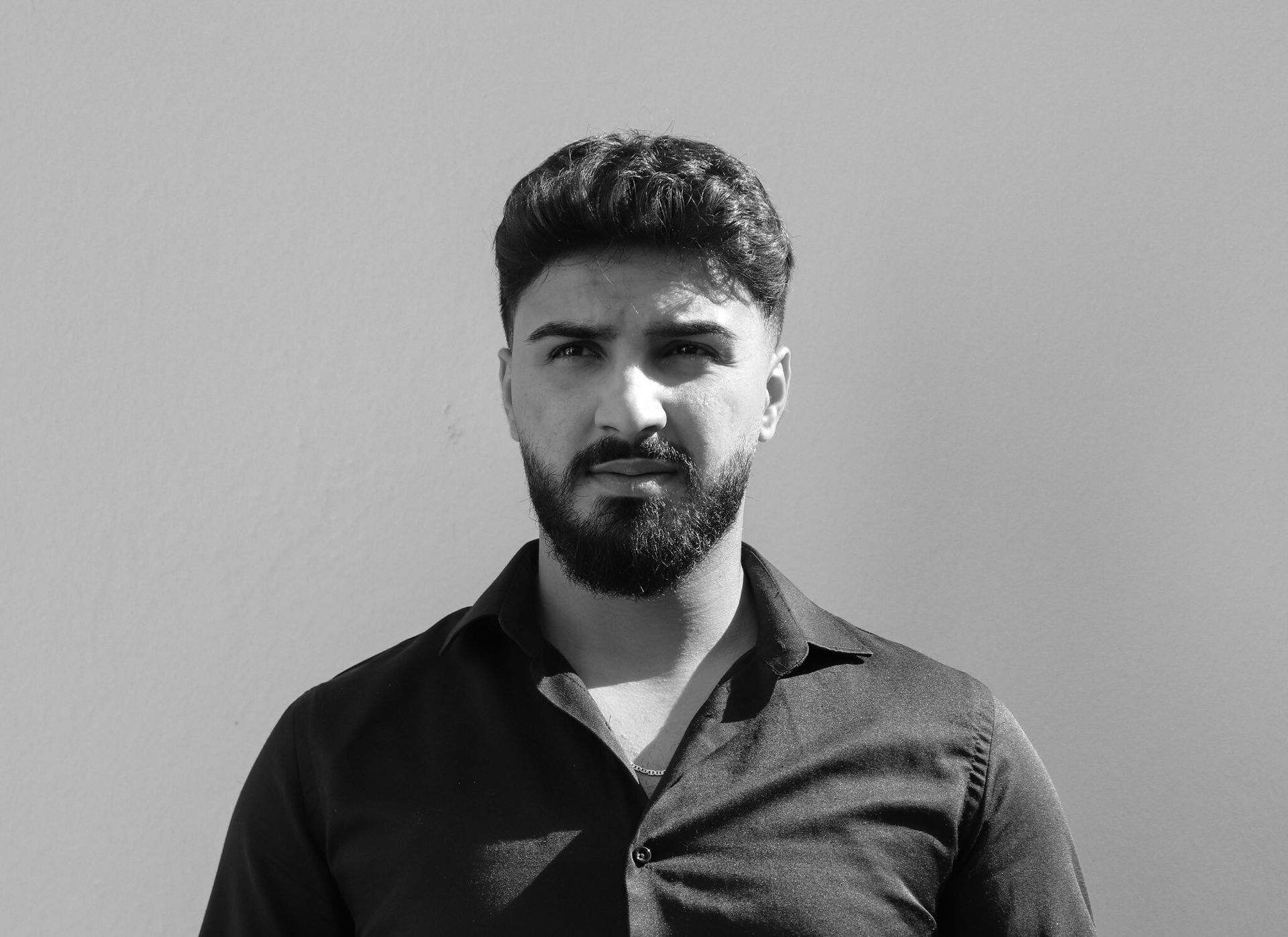 Mann mit dunklem Haar und Bart vor hellem Hintergrund, trägt ein dunkles Button-Down-Hemd und blickt in die Kamera.
