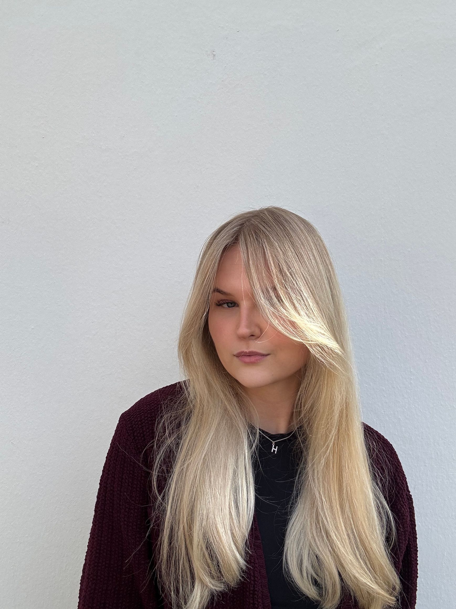 Blonde Frau mit langen Haaren und Pony in einer burgunderfarbenen Strickjacke vor einer weißen Wand.
