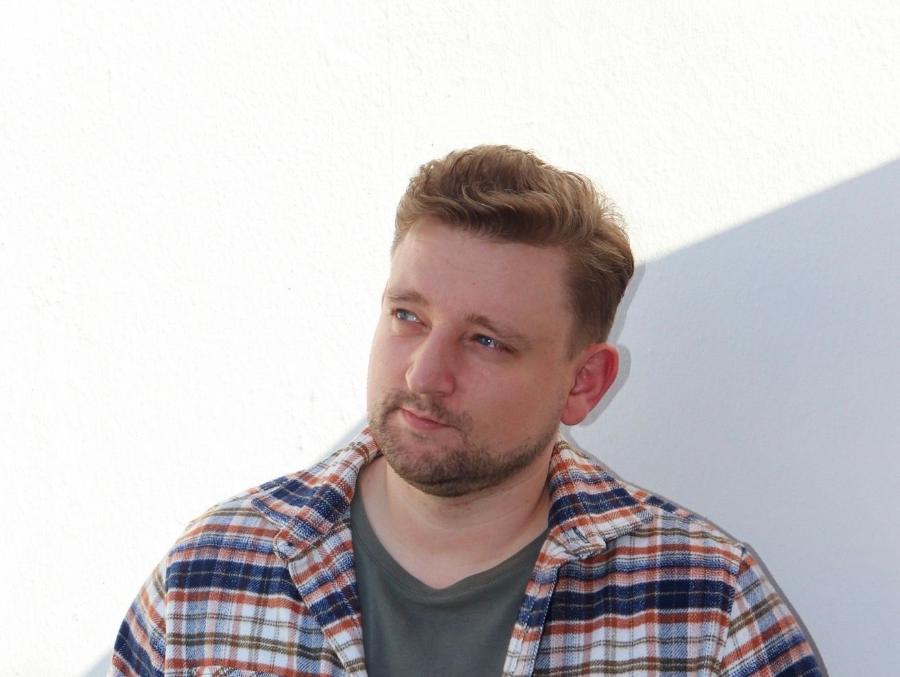 Mann mit hellem Haar und Bart, trägt ein kariertes Hemd, blickt zur Seite und lehnt an einer weißen Wand.