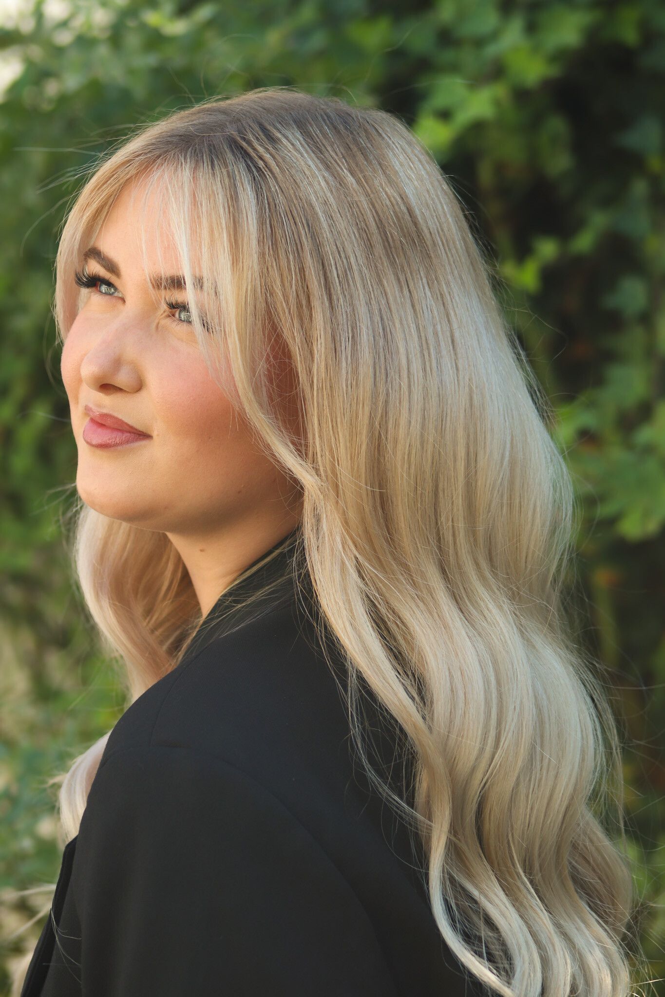 Blonde Frau mit leichtem Make-up, Blick zur Seite, dunkle Jacke, draußen im Grünen.