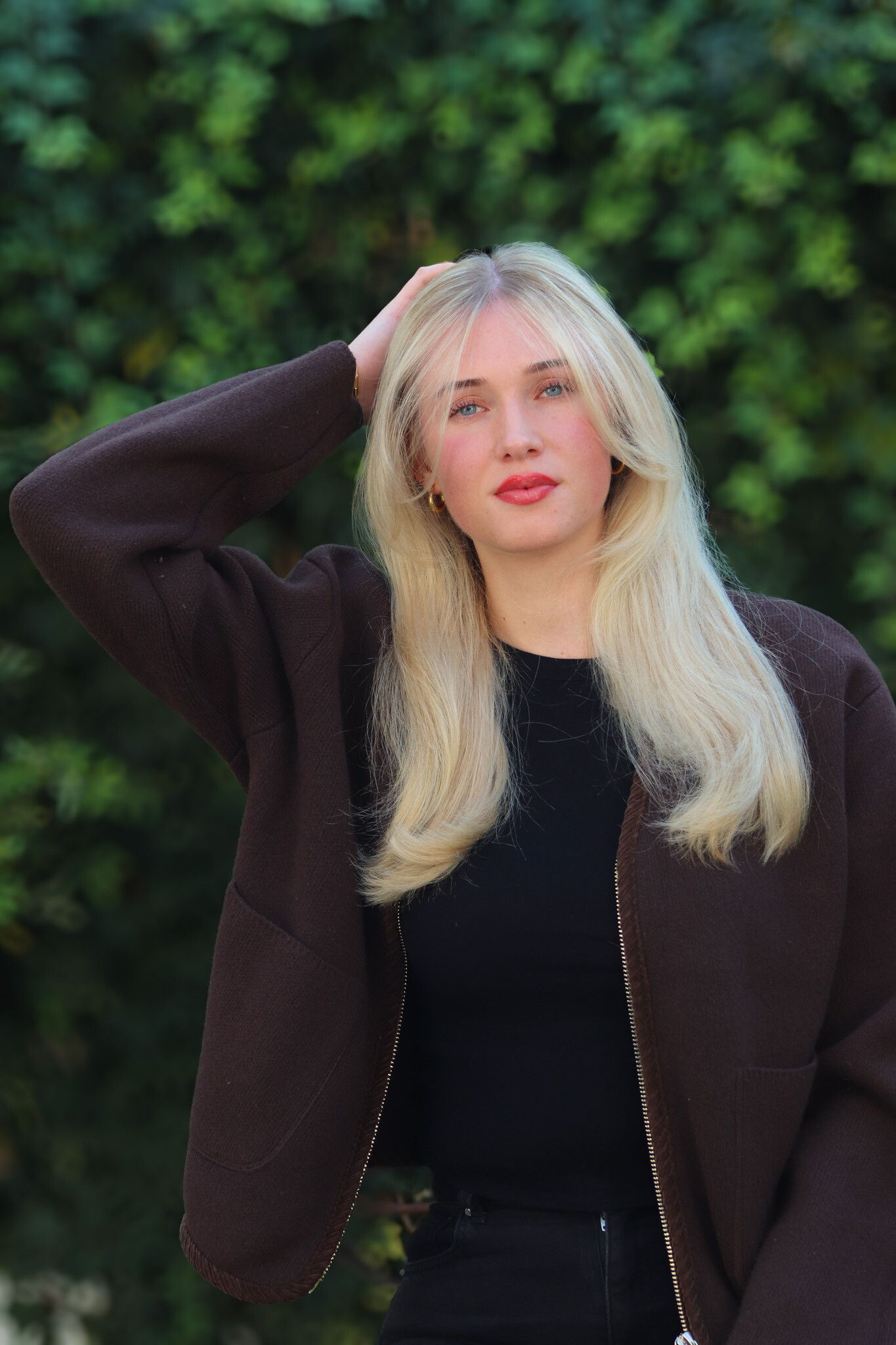 Blonde Frau mit der Hand im Haar, trägt eine dunkle Jacke und ein schwarzes Oberteil und steht vor grünem Laub.