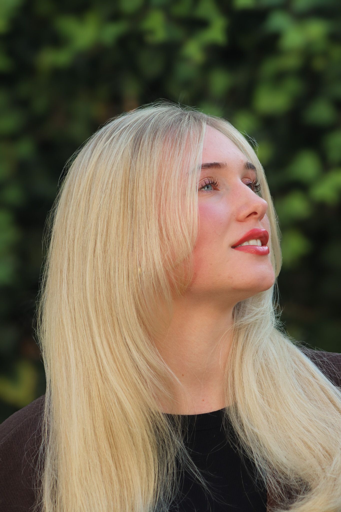 Blonde Frau blickt lächelnd nach oben vor verschwommenem grünem Laub.