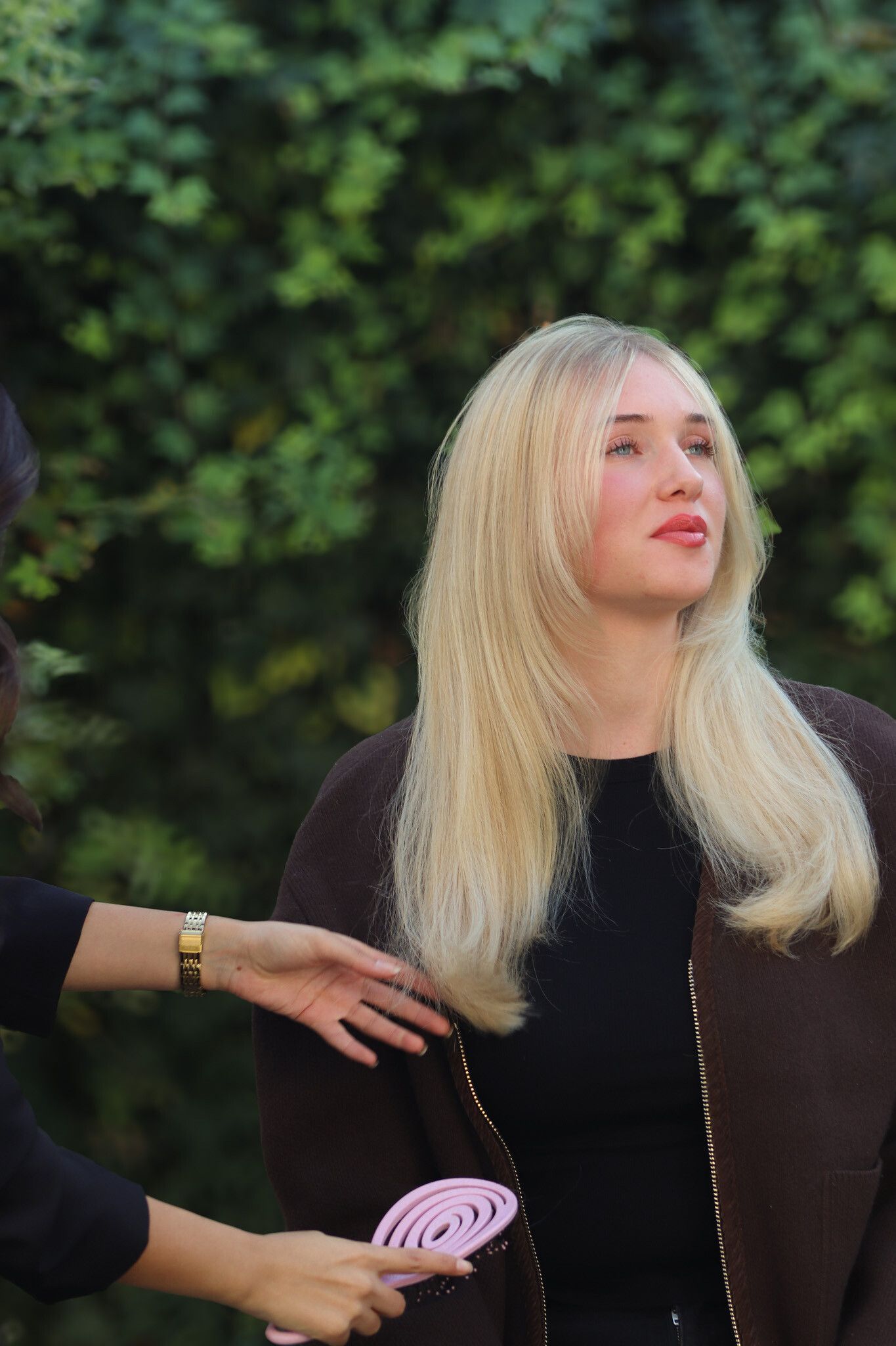Blonde Frau in einem braunen Mantel erhält Hilfe mit einem rosa Fächer. Im Freien, grüner Laubhintergrund.