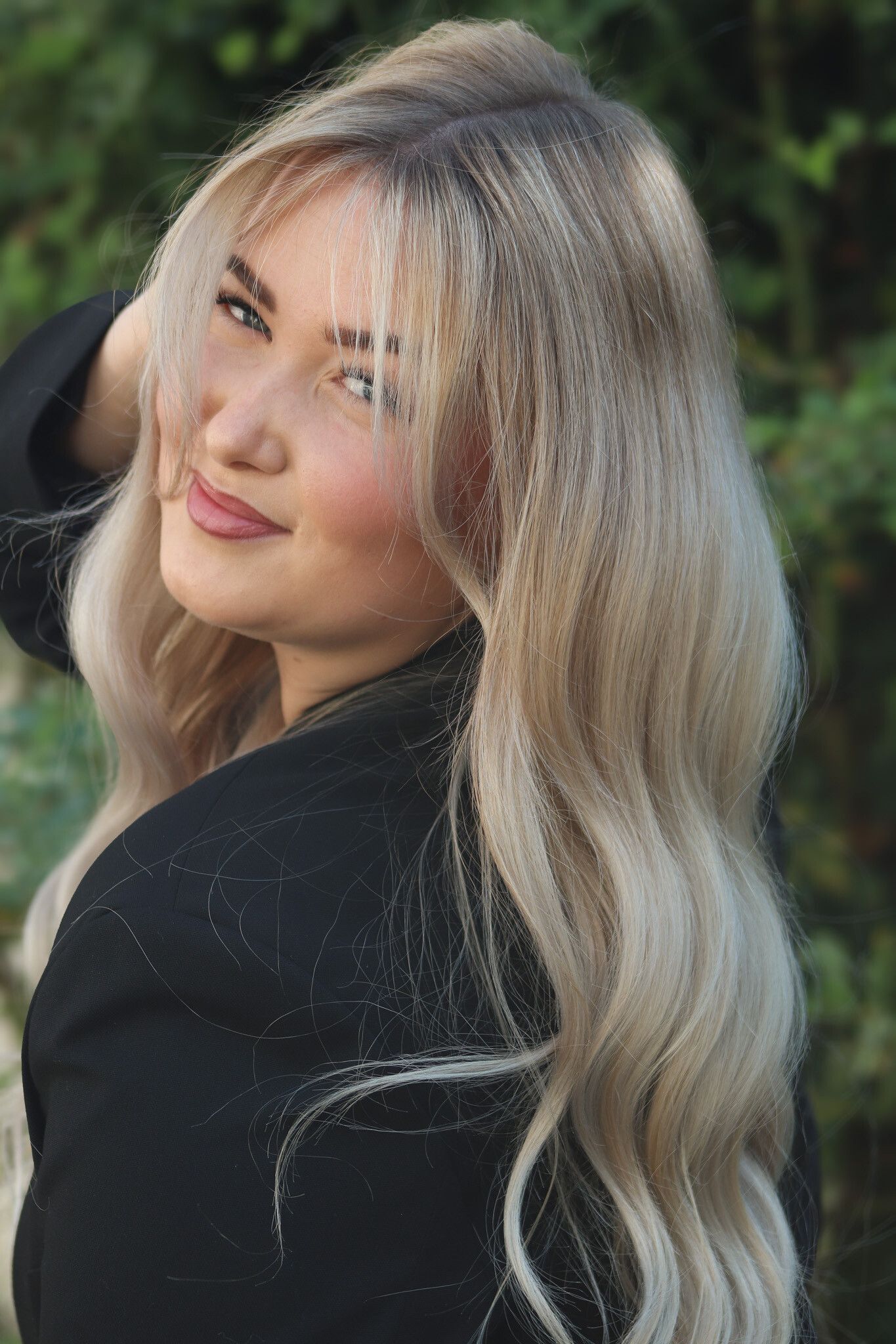 Blonde Frau lächelt und blickt über ihre Schulter. Im Freien, trägt eine schwarze Jacke, die Hand im Haar.