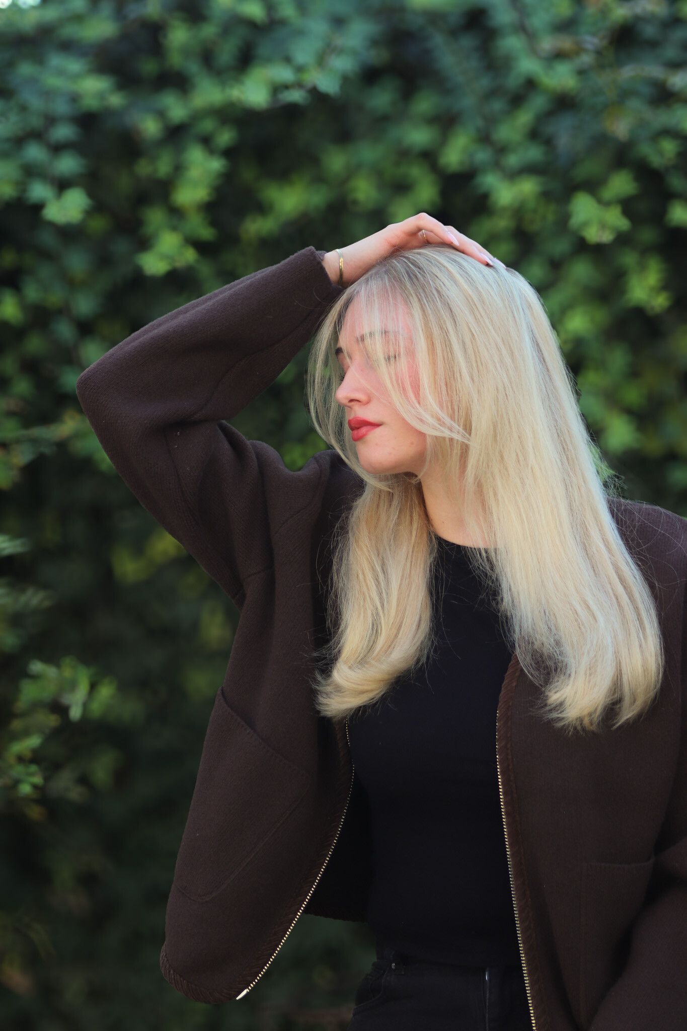 Blonde Frau in dunkler Jacke, Hand auf dem Kopf, rote Lippen, vor grünem Laubhintergrund.