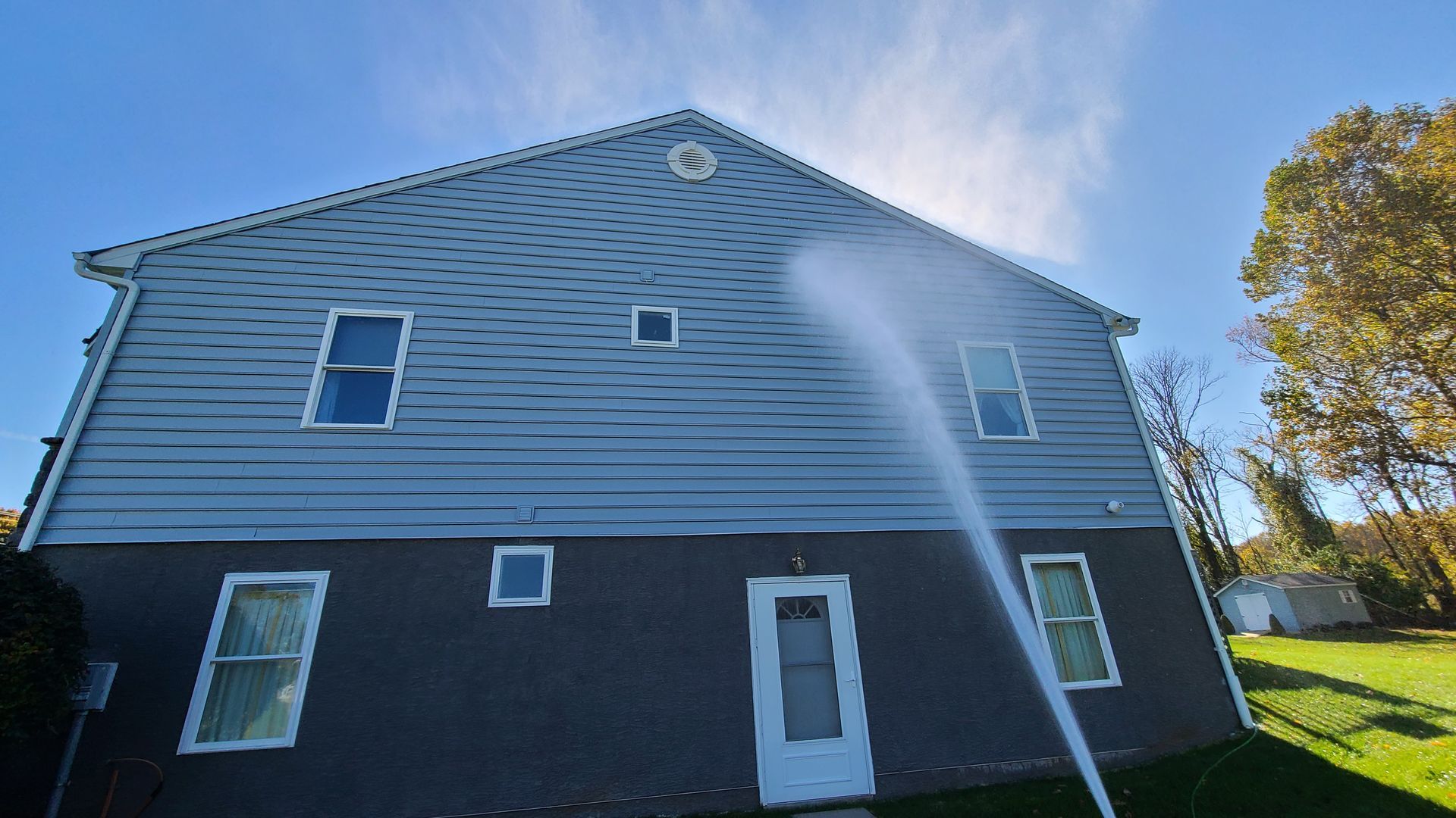Pressure washing a two-story house with blue siding on a sunny day.