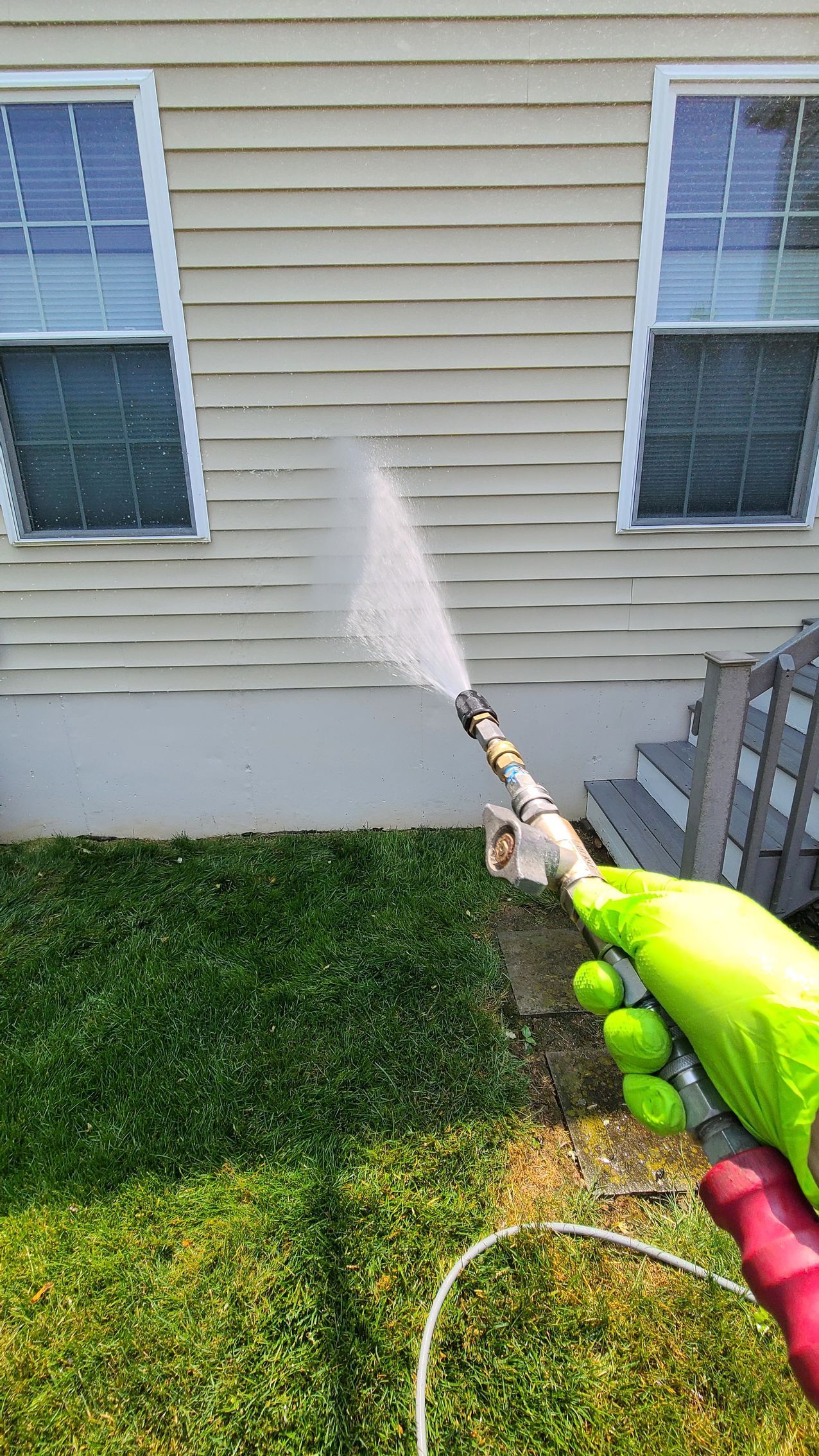 Person power washing siding on a house with a pressure washer; green lawn visible.