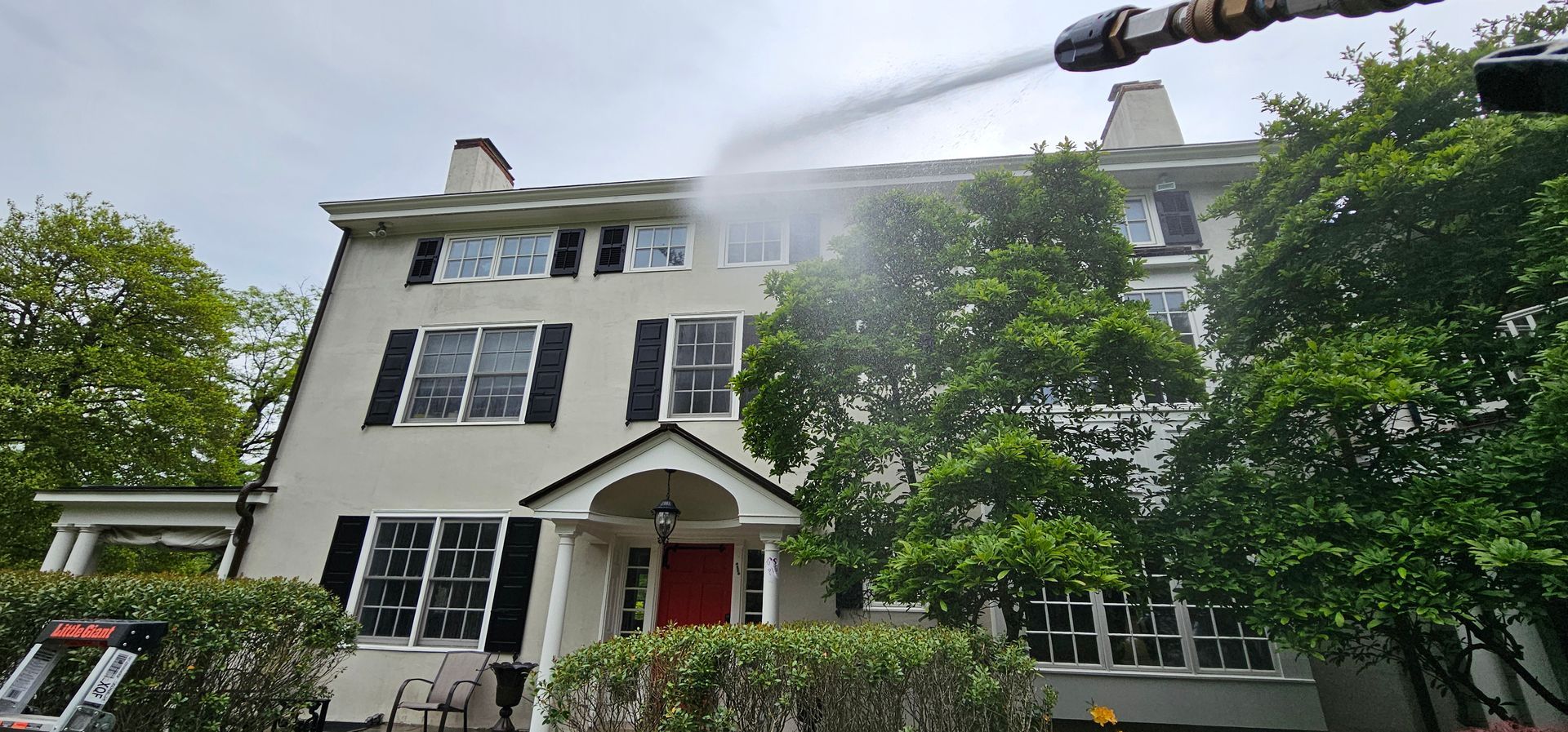 Pressure washing a large, off-white house with black shutters, trees, and a red door.