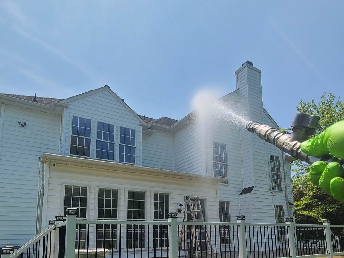 Person pressure washing a white house under a blue sky.