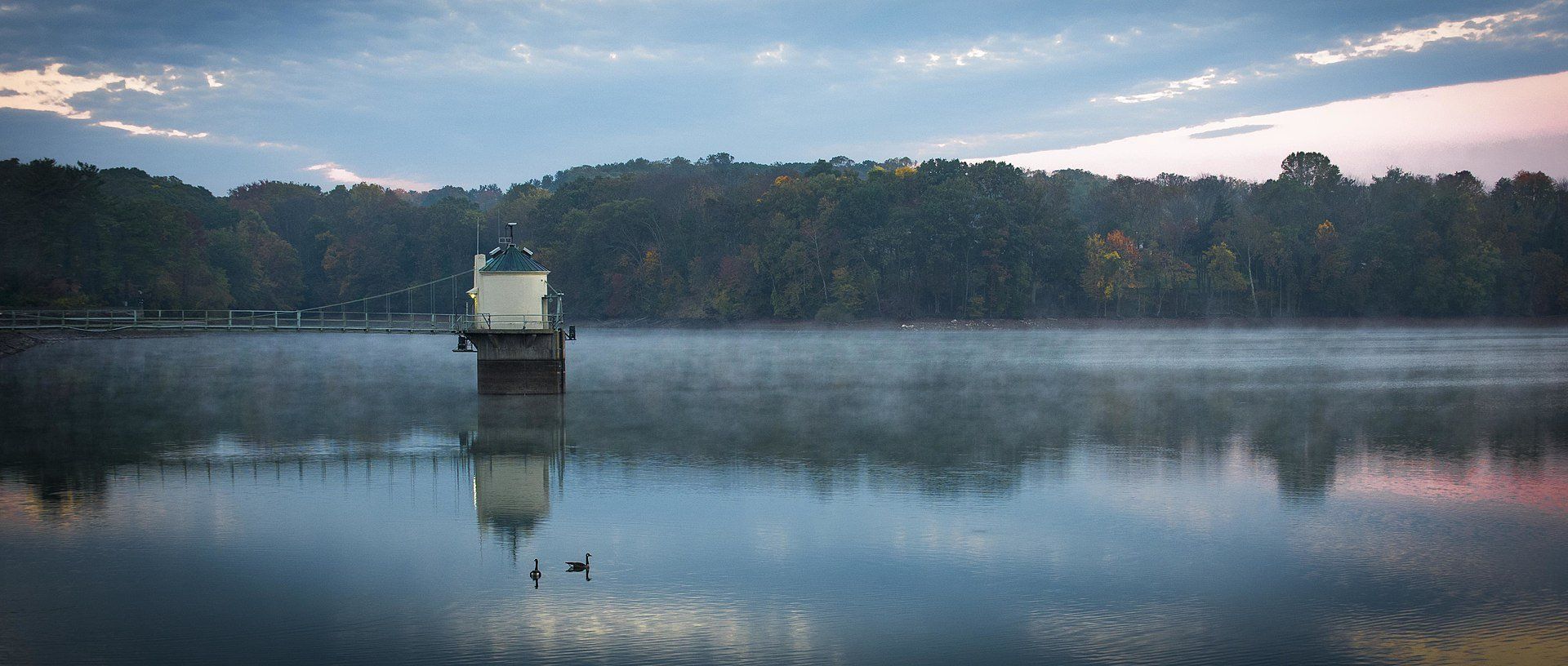 A tranquil lake scene at dusk. A structure is in the water, with tree-lined shore and colorful sky.
