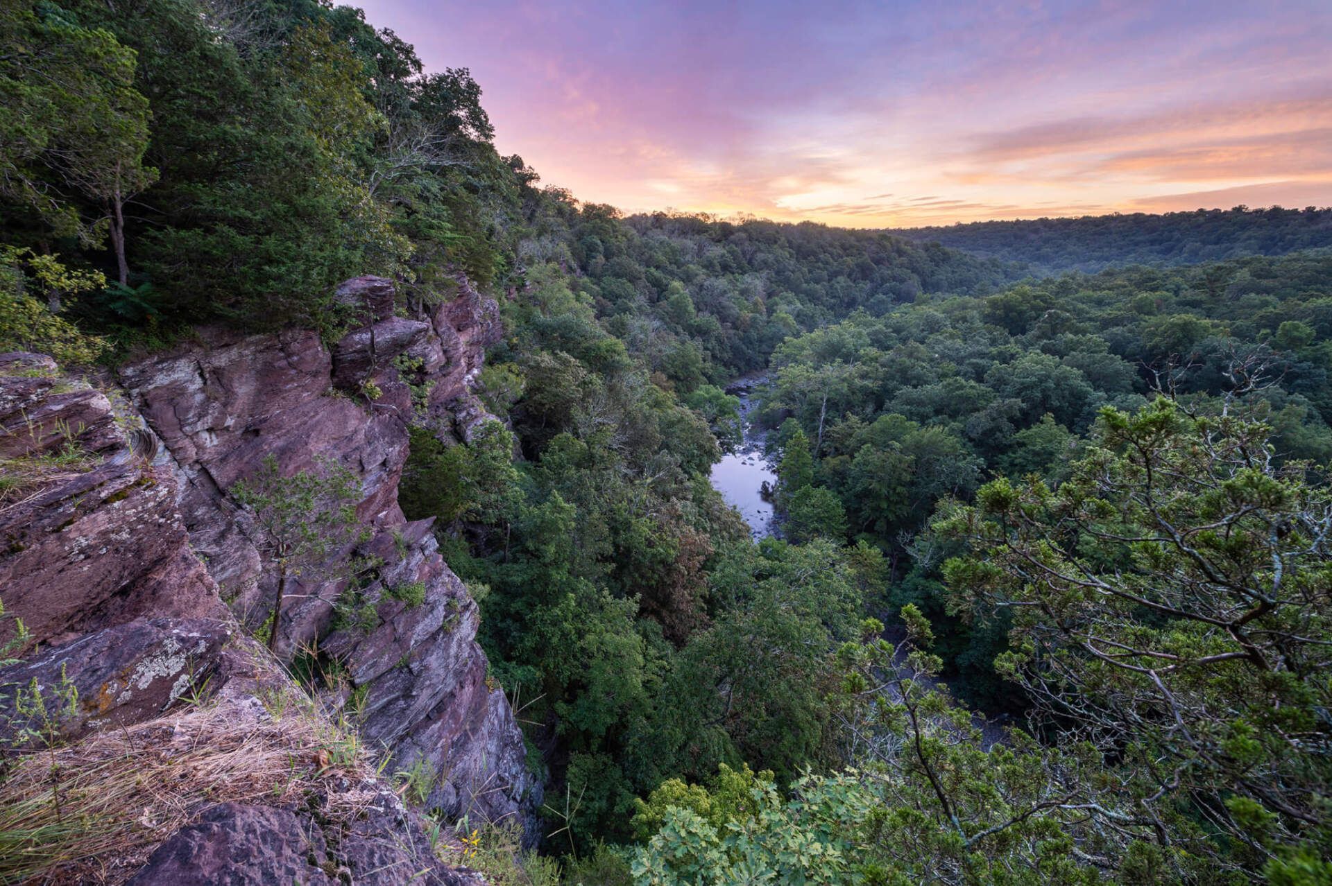 Cliff overlooking a valley filled with lush green trees and a winding river; sunset sky.