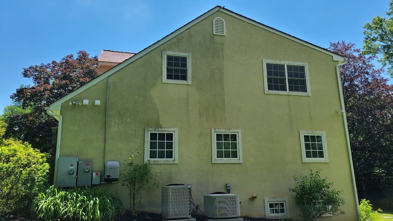 Back of a two-story beige house with multiple windows and visible algae growth on the stucco exterior under a blue sky.