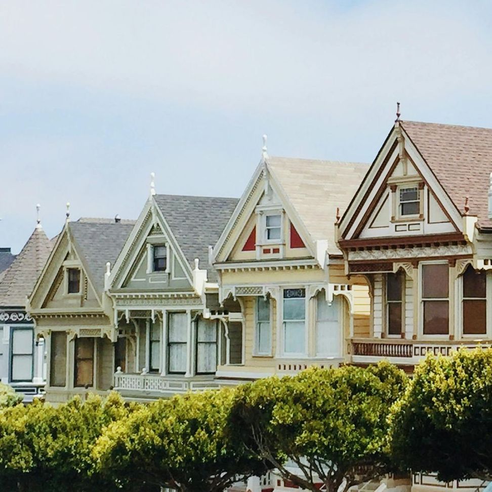 Victorian houses, colorful facades, and decorative trim, line a street, with green bushes below.
