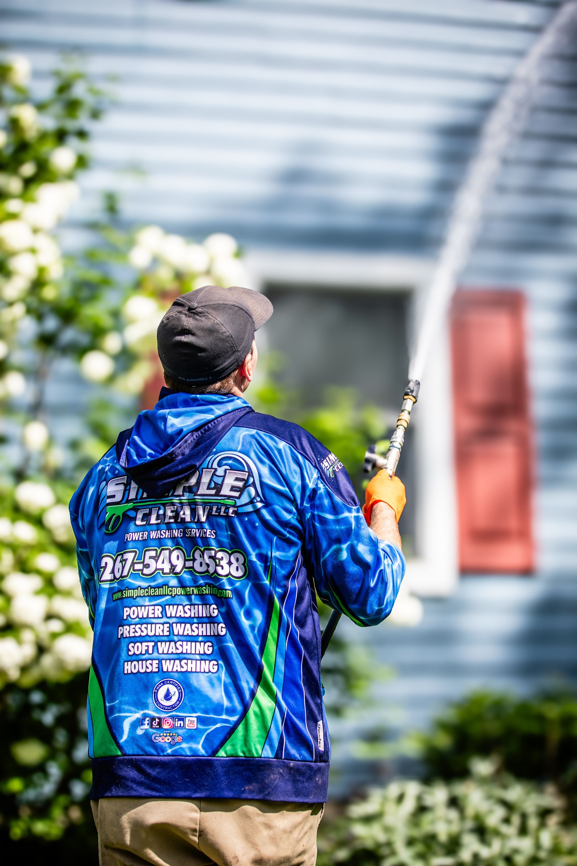 Person pressure washing the exterior of a blue house; sporting a blue sweatshirt, gloves, and a cap.