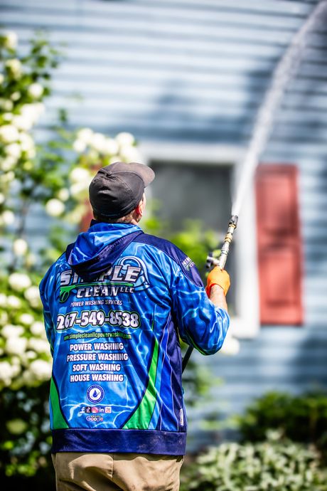 Person in blue shirt power washing a house; outdoor daytime.