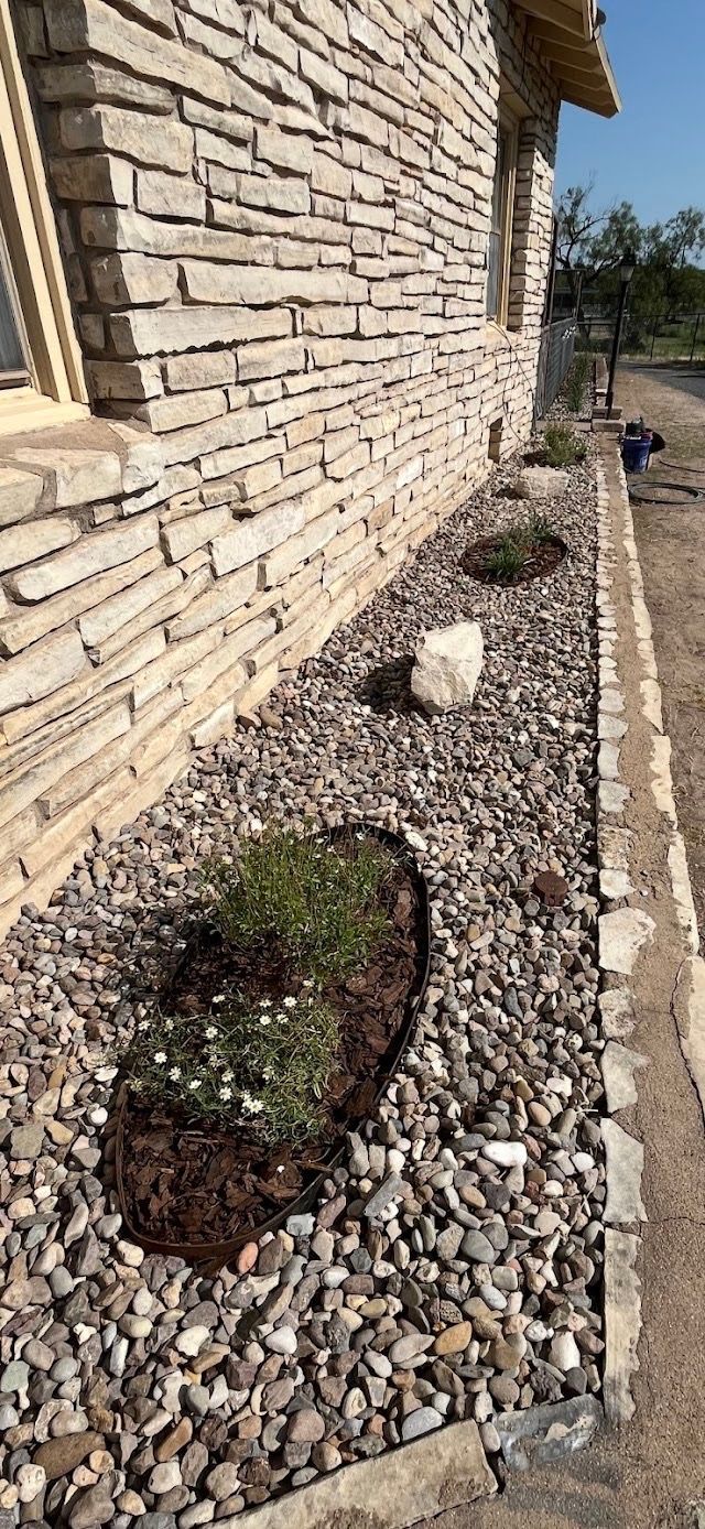 Stone-clad building with a gravel-covered flower bed. A rock, and several small plants are visible.