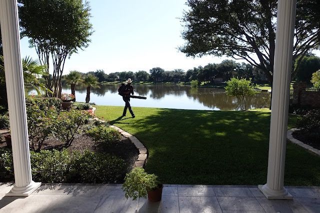 Person using a leaf blower on green lawn by a lake, viewed from a porch with columns.