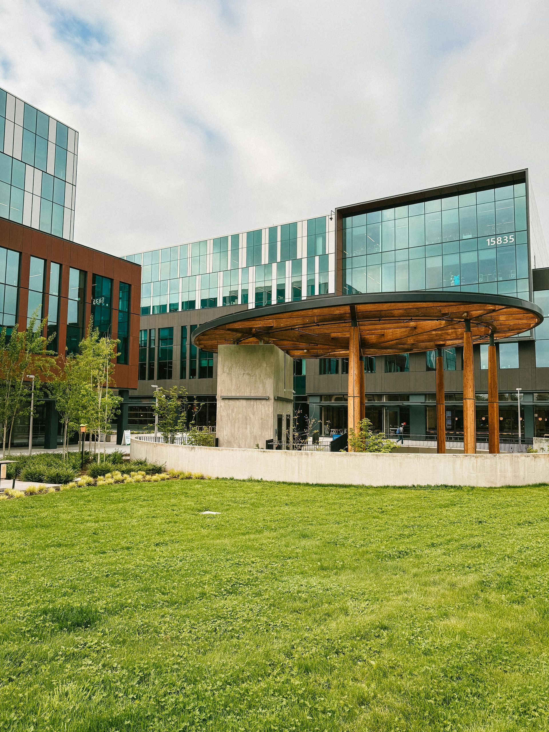 Lush green lawn in front of modern buildings with glass windows. A wooden canopy offers shade.