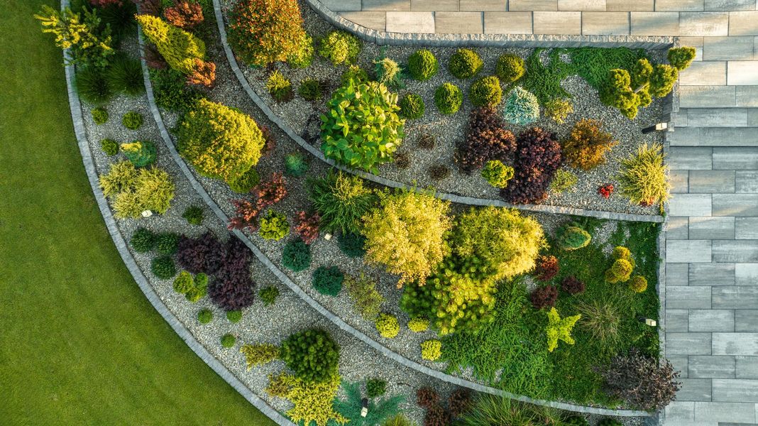 Overhead view of a tiered garden bed with colorful plants, gravel, and stone bordering. Green lawn on the left.