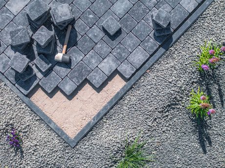 Pathway construction with dark gray pavers, a hammer, and gravel base.