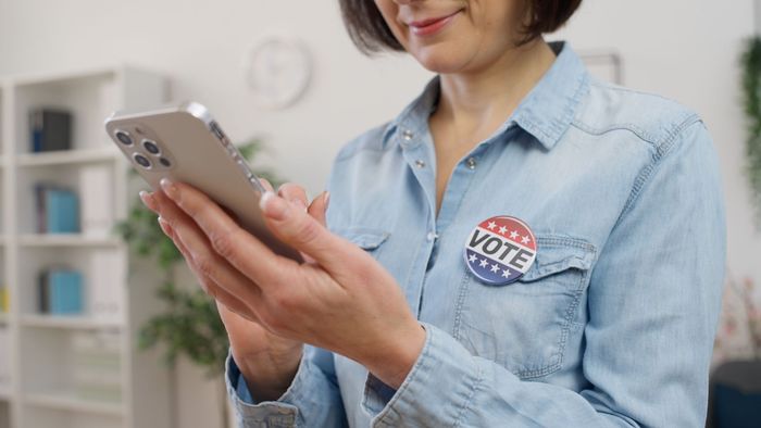 A woman with a vote button on her shirt is using a cell phone.