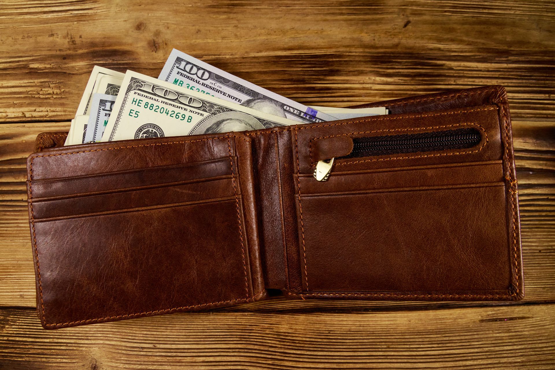 Brown leather wallet open with cash visible on a wooden surface.