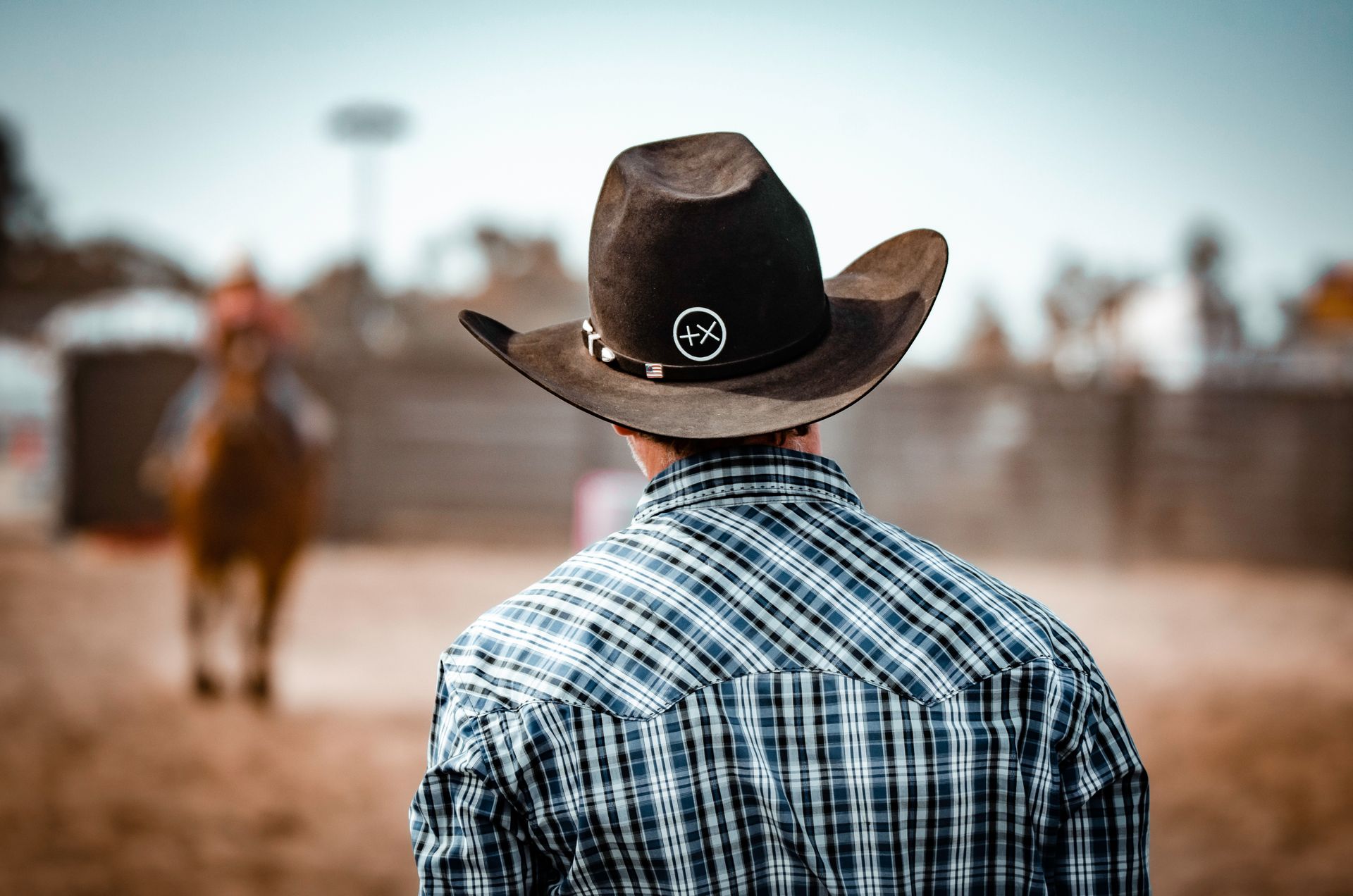 Person in cowboy hat watches rodeo event. Plaid shirt, brown hat with emblem.