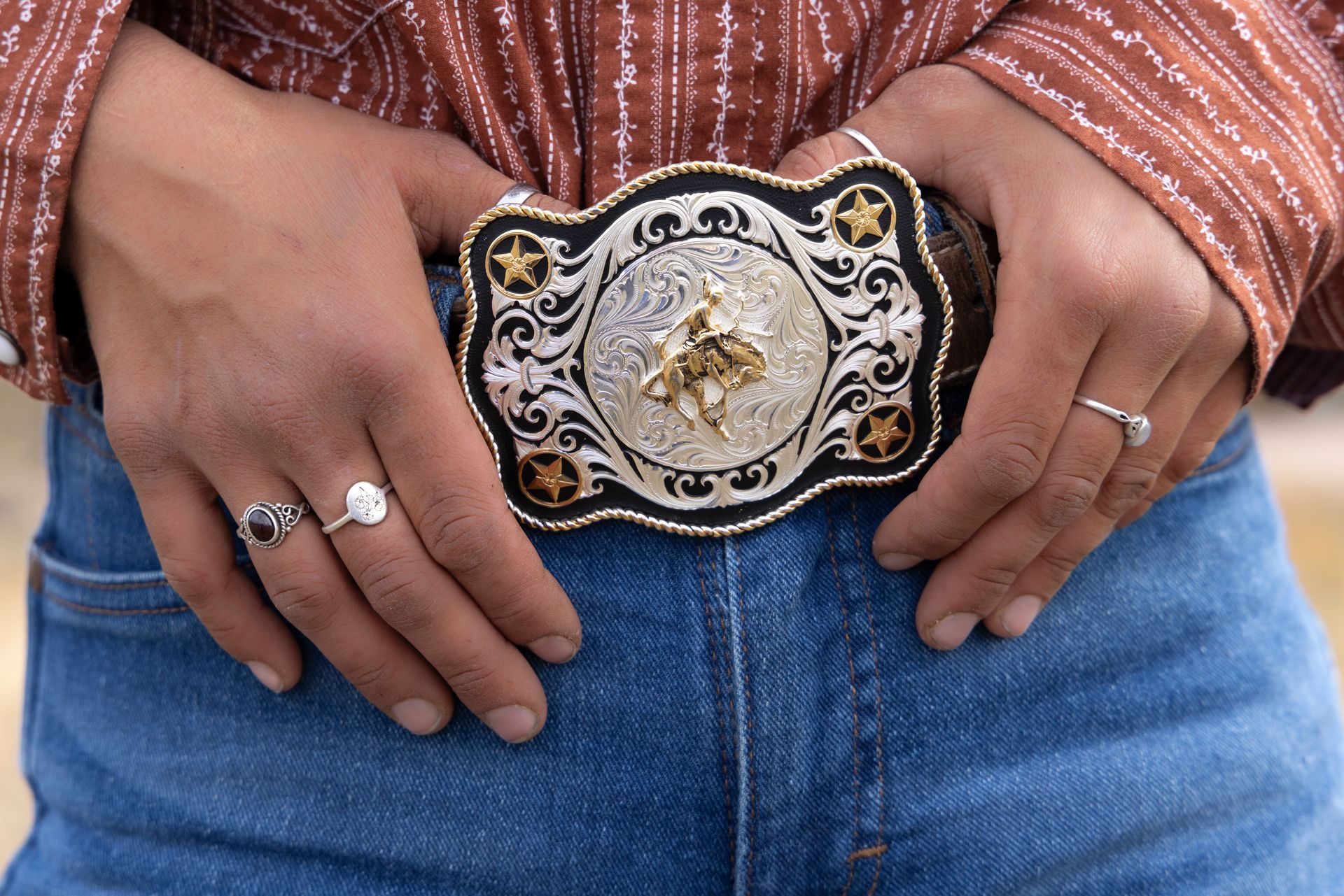 Person wearing jeans with a large, ornate silver and gold belt buckle and rings.