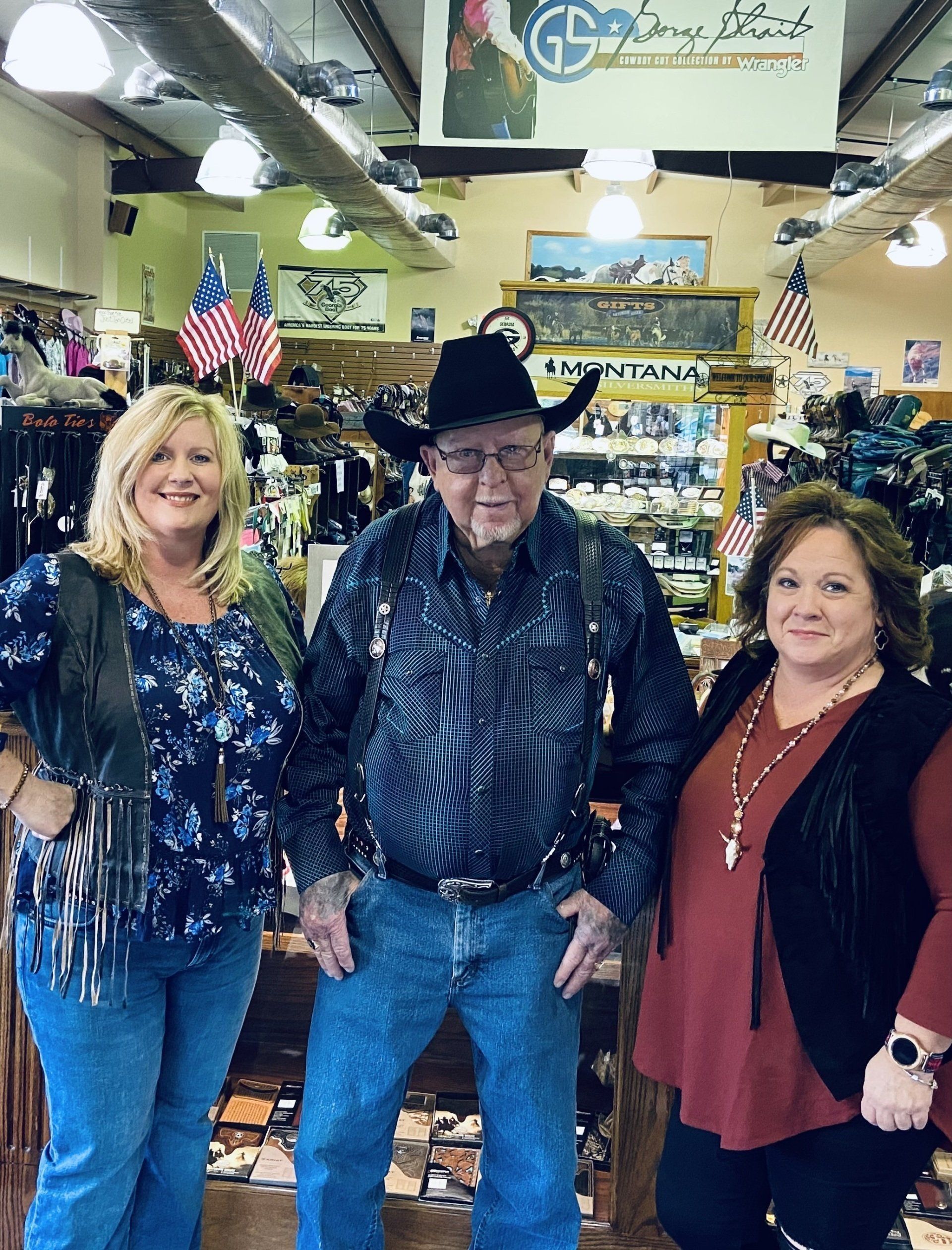 Three people posing inside a store. Man in cowboy hat and suspenders flanked by women wearing vests, all smiling.